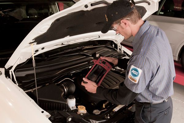 Mechanic in uniform using diagnostic tool under the hood of a white car in a garage.