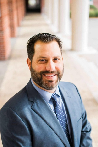 Man in a blue suit and tie, smiling, outdoors near brick columns.