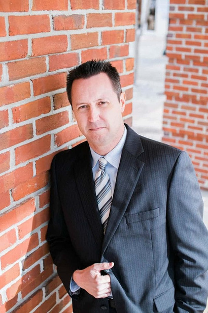 Man in a suit leaning against a brick wall, smiling slightly.