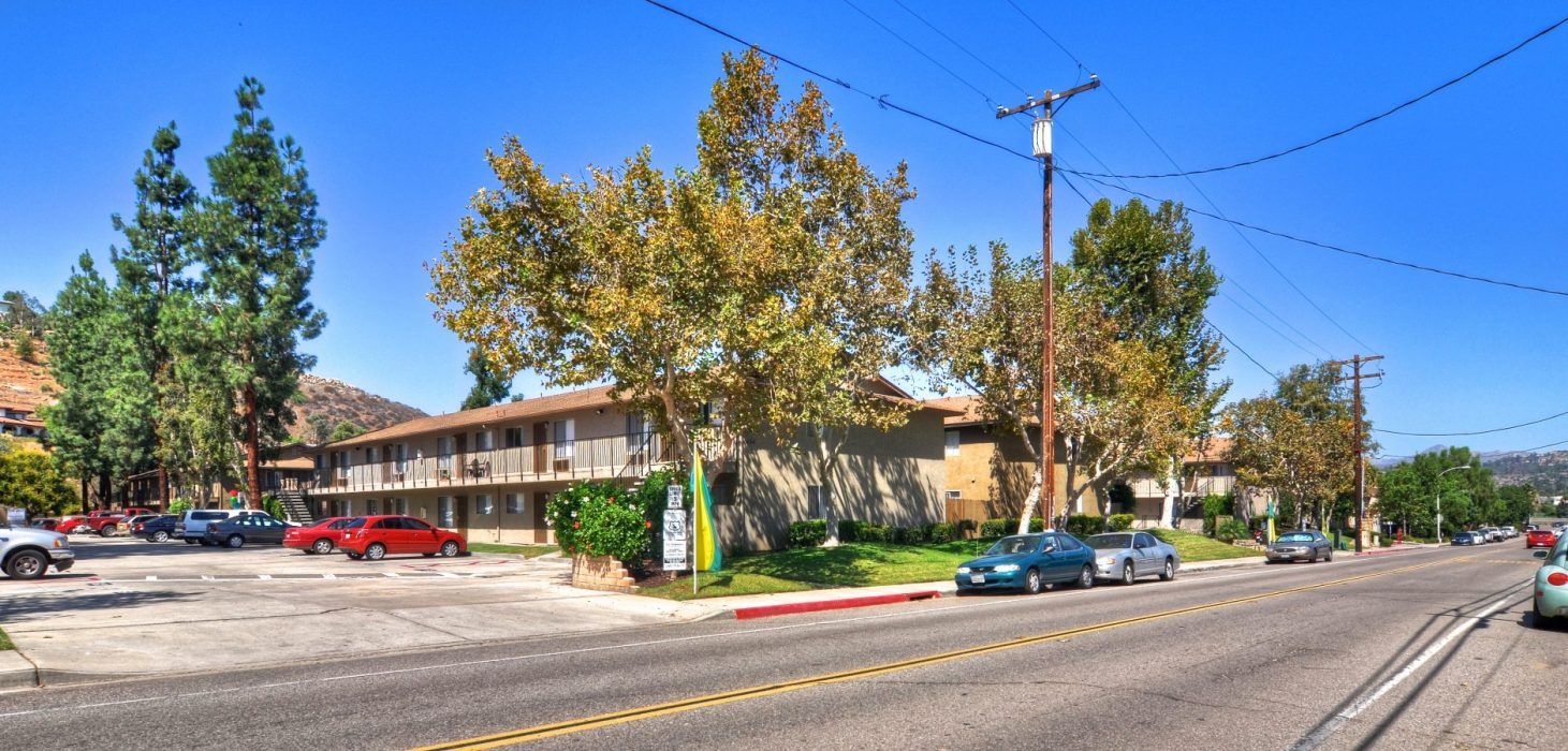 Sunny street corner with apartment building, parked cars, trees, and clear blue sky