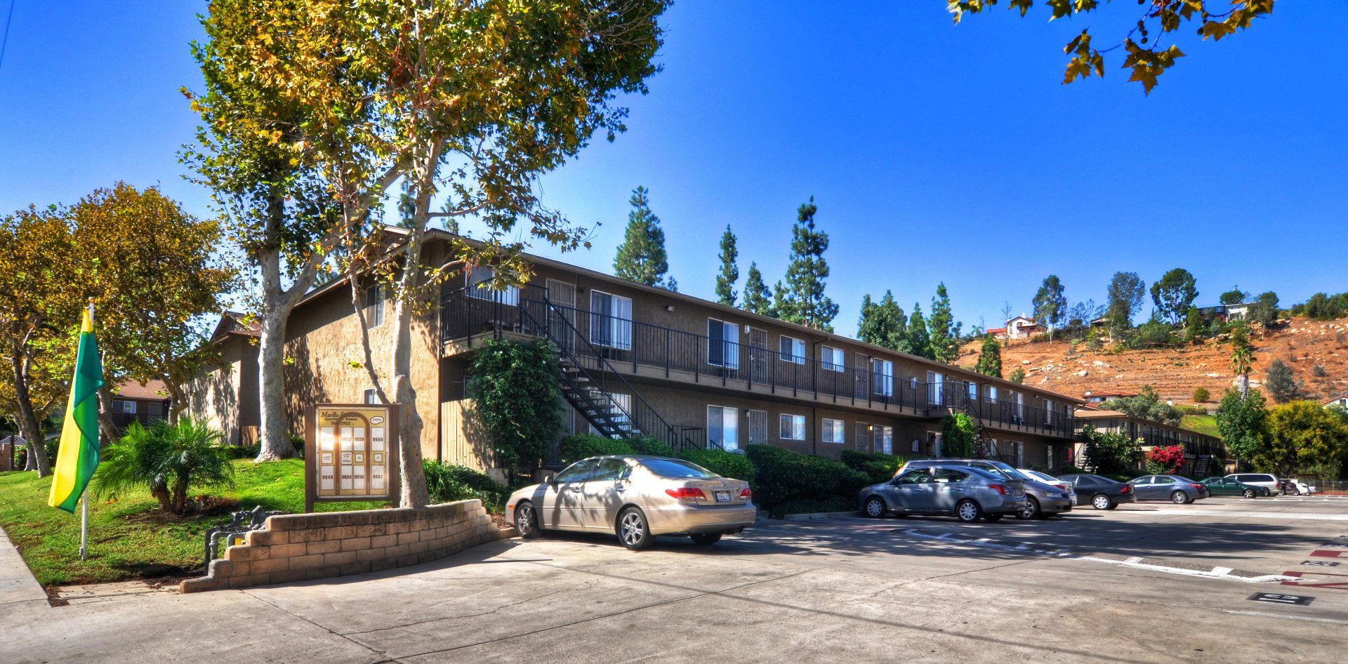 Two-story apartment building with parked cars and trees in a sunny lot