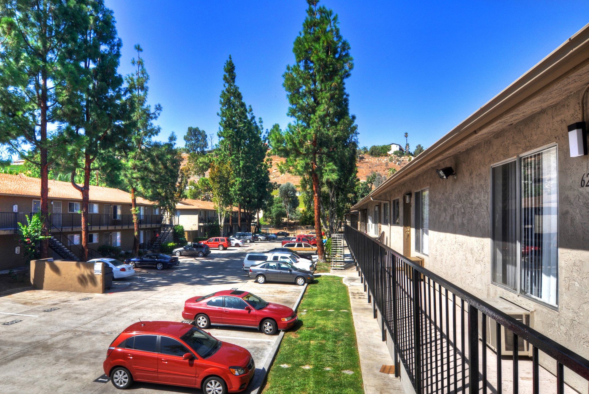 Apartment parking lot with red cars, trees, and a walkway beside a building on a sunny day