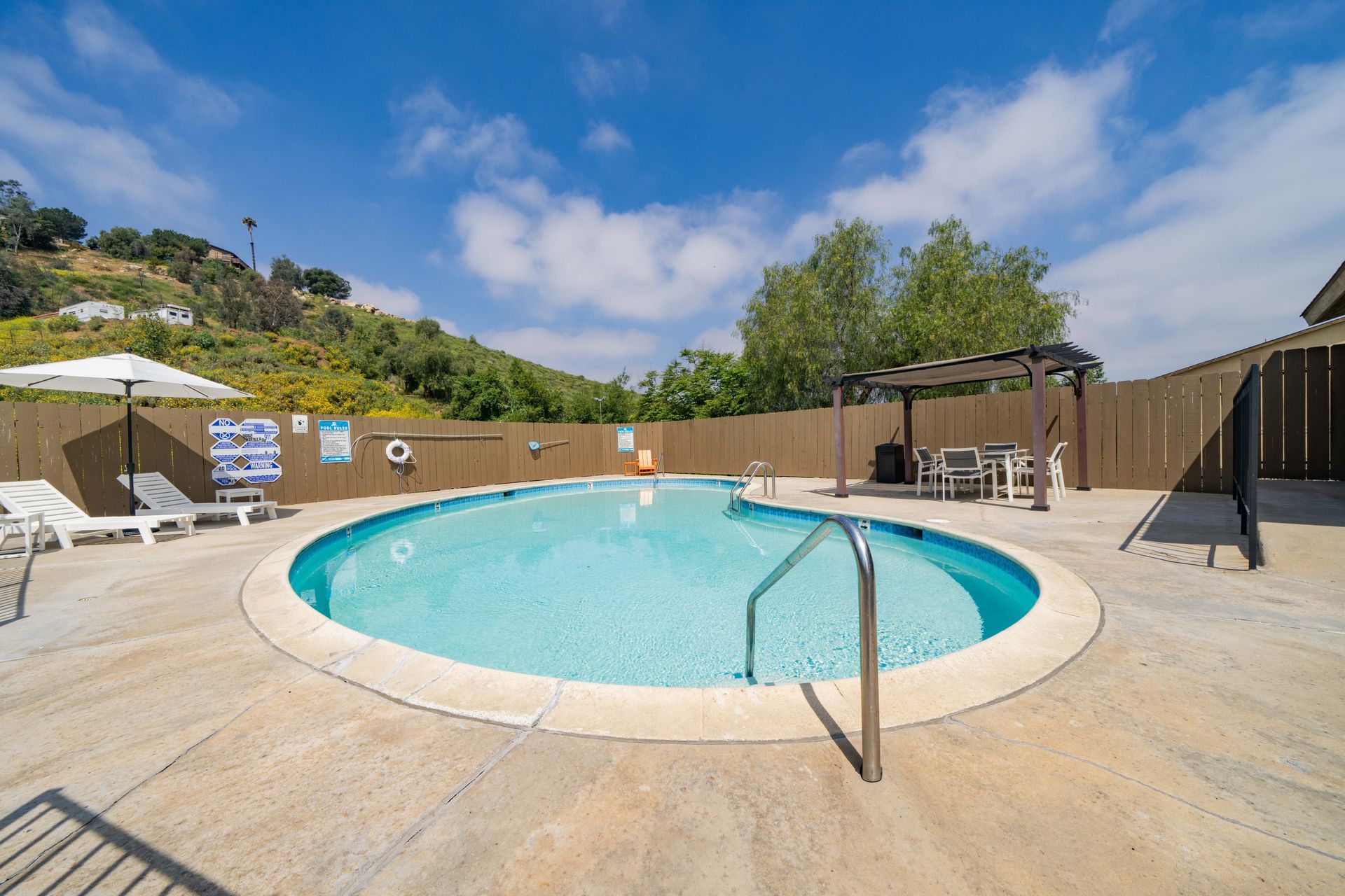 Outdoor swimming pool with lounge chairs, wooden fence, and hillside under a blue sky.