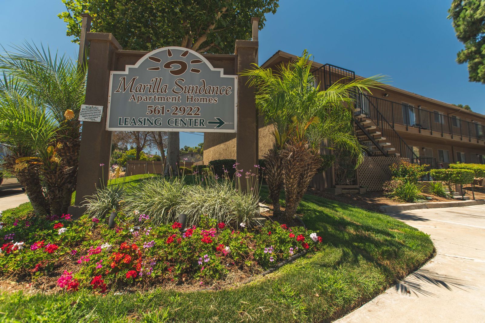 Historic building entrance with a decorative sign, bright flowers, and a sunlit walkway.