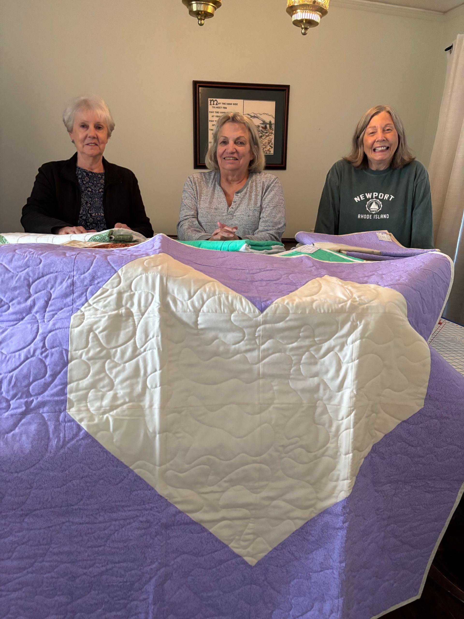 Three older women are sitting at a table with a heart shaped quilt.