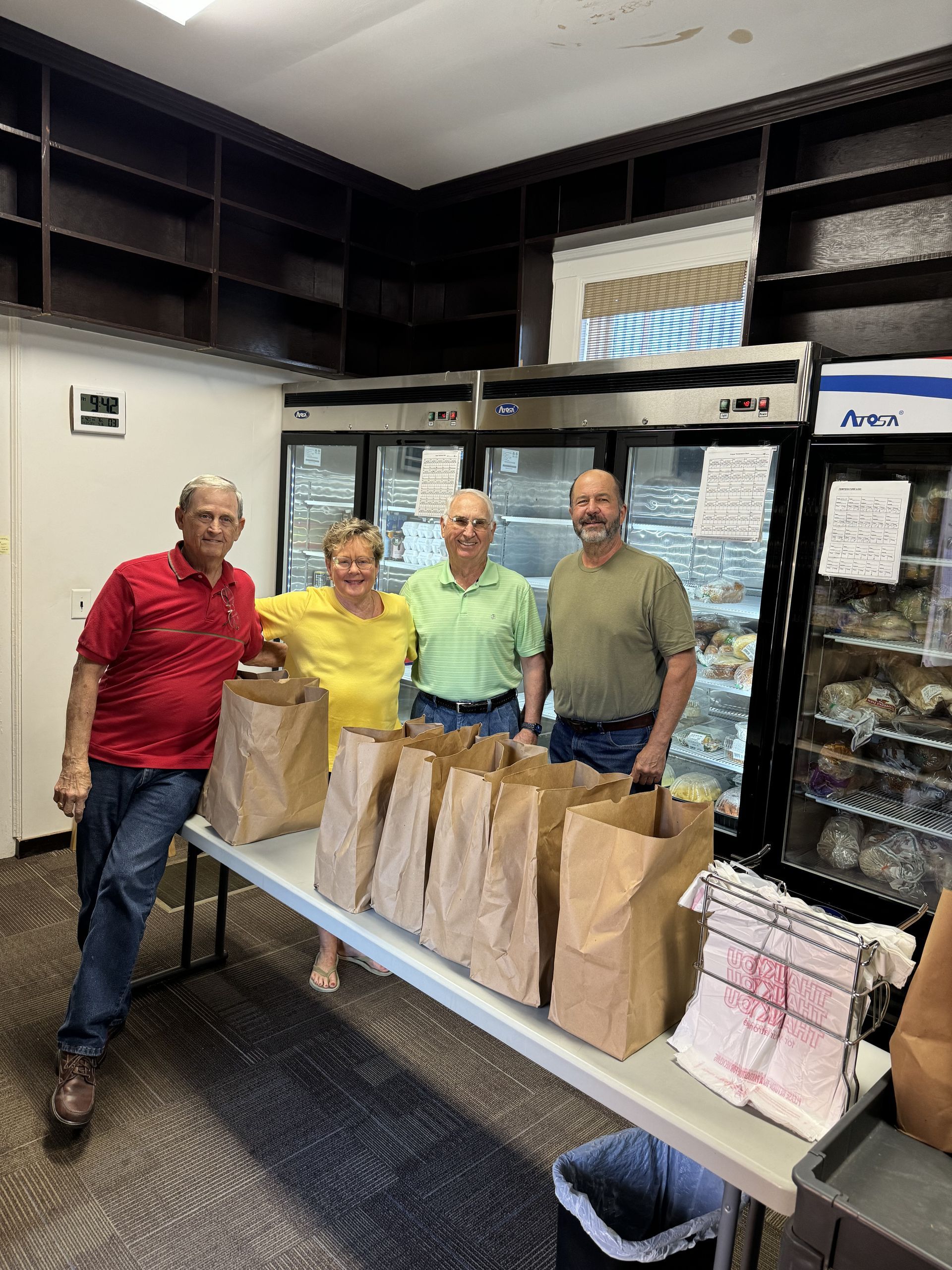 A group of 4 older adults standing around a table that has brown paper bags on it.