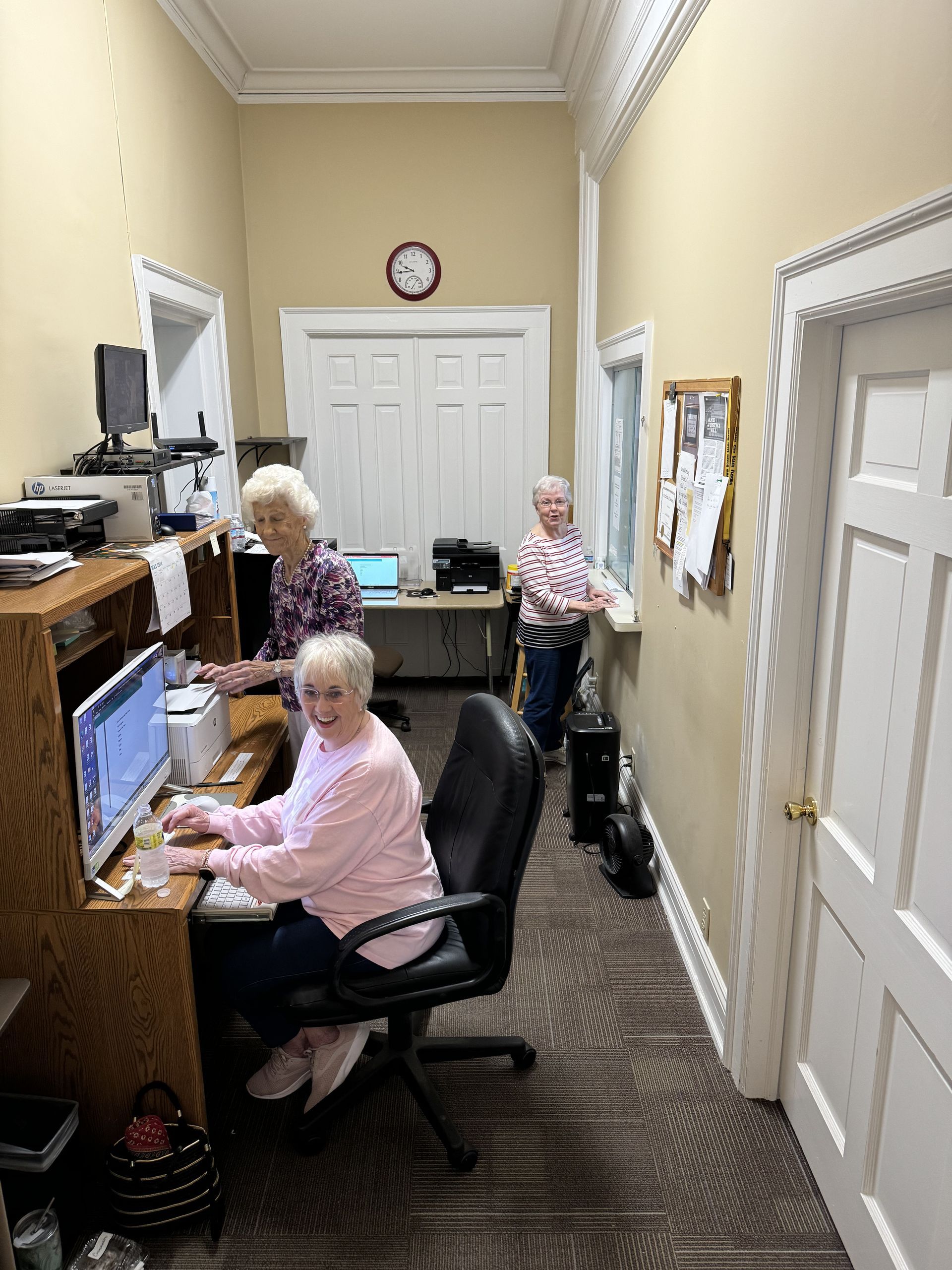 Three older women are sitting at desks in an office using computers.