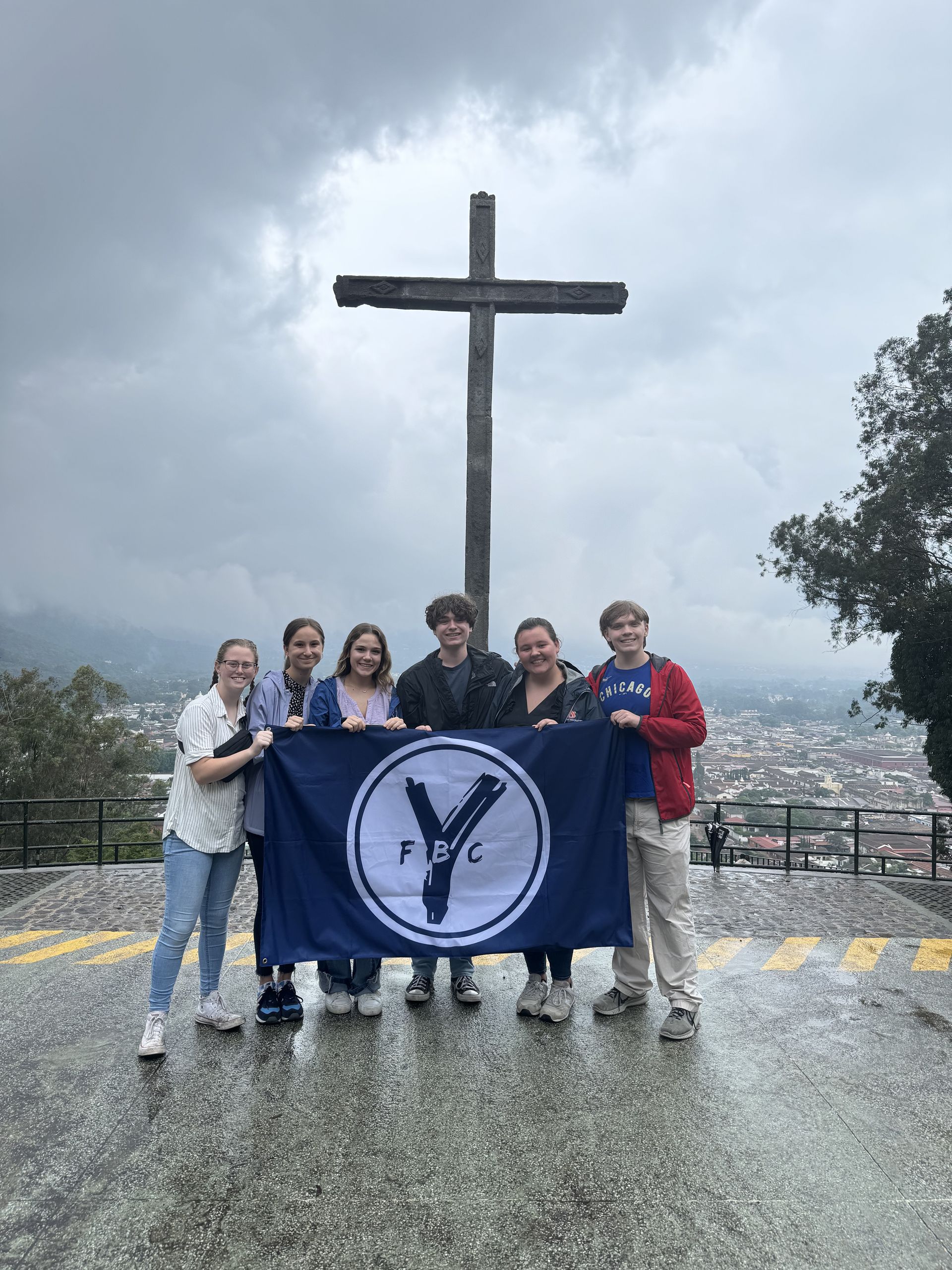 Youth Group students are standing in front of a cross holding a First Baptist Church Youth flag in Guatemala.