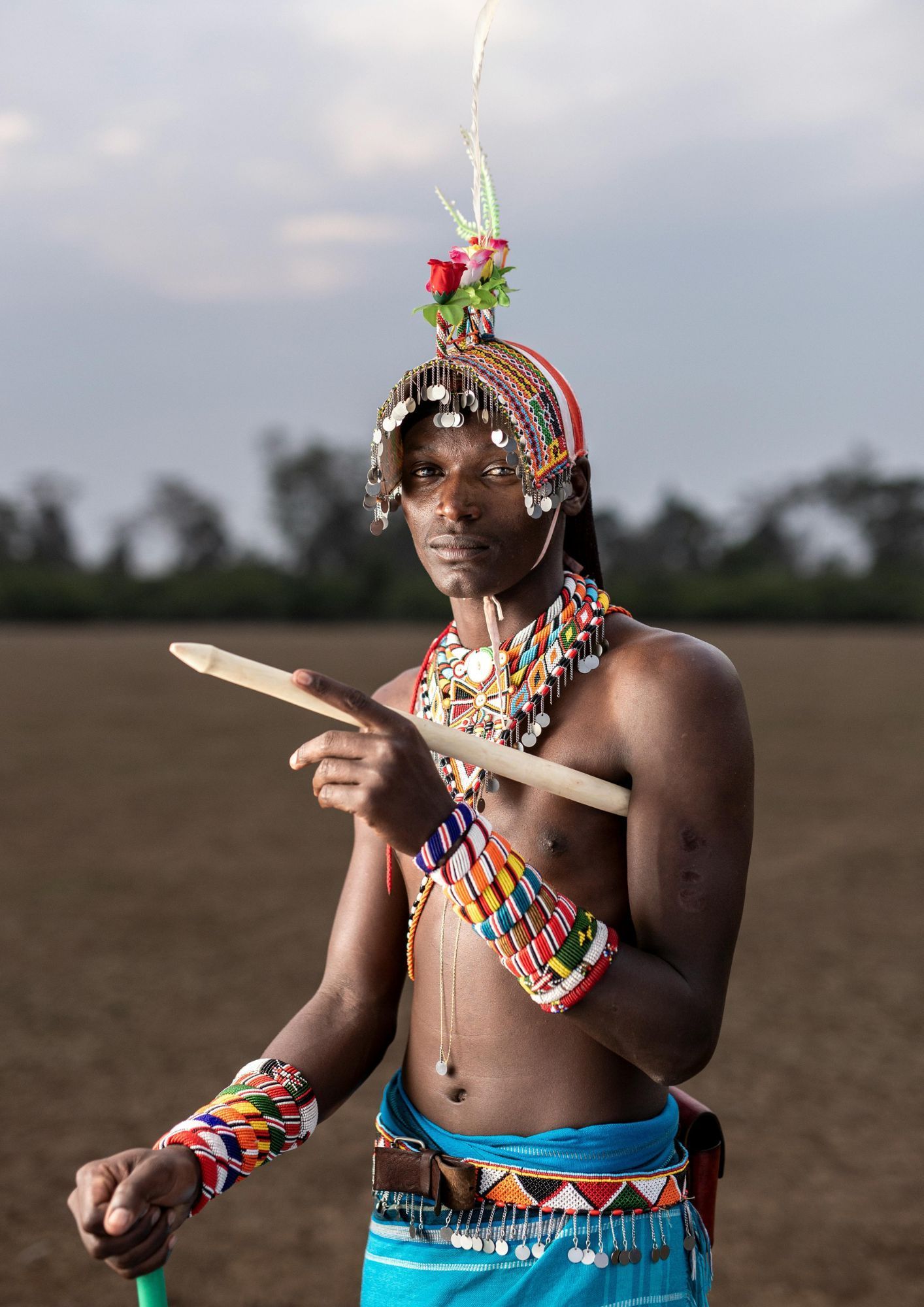 Kenyan man in traditional attire, pointing a wooden stick, beaded jewelry, open field.