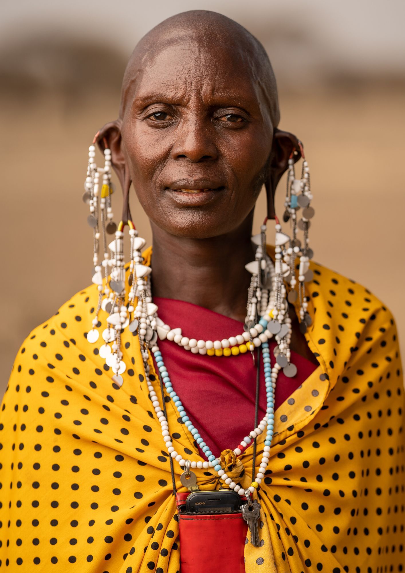 Maasai woman wearing large earrings and beaded necklaces, draped in a yellow shawl with black polka dots.