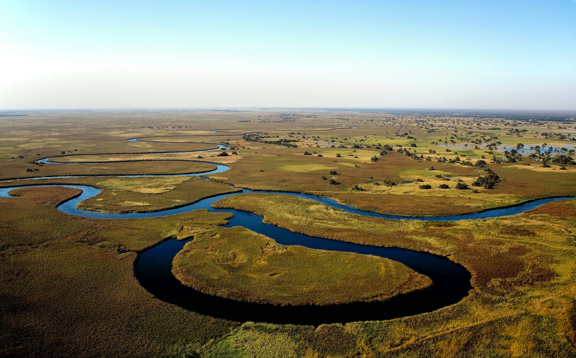 Aerial view of a winding river through marshy, green-brown terrain under a clear blue sky.