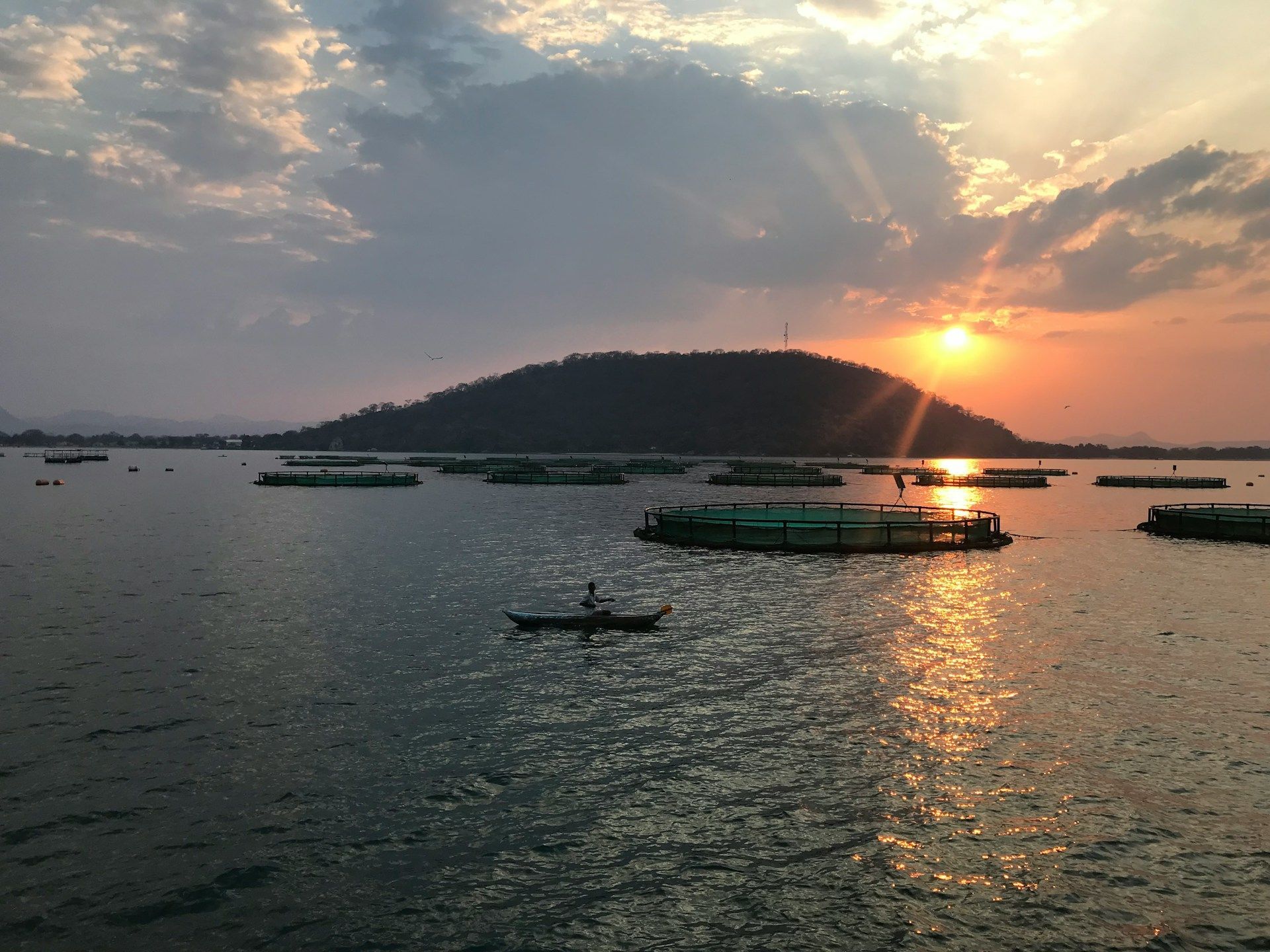 Sunset over a lake with floating structures, a person in a boat, and a silhouetted hill.