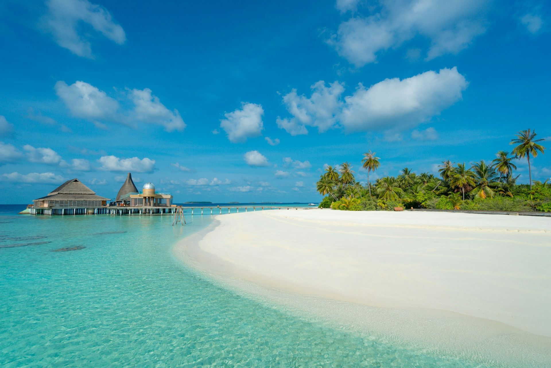 White sandy beach with crystal clear turquoise water, overwater huts, and blue sky with fluffy clouds.