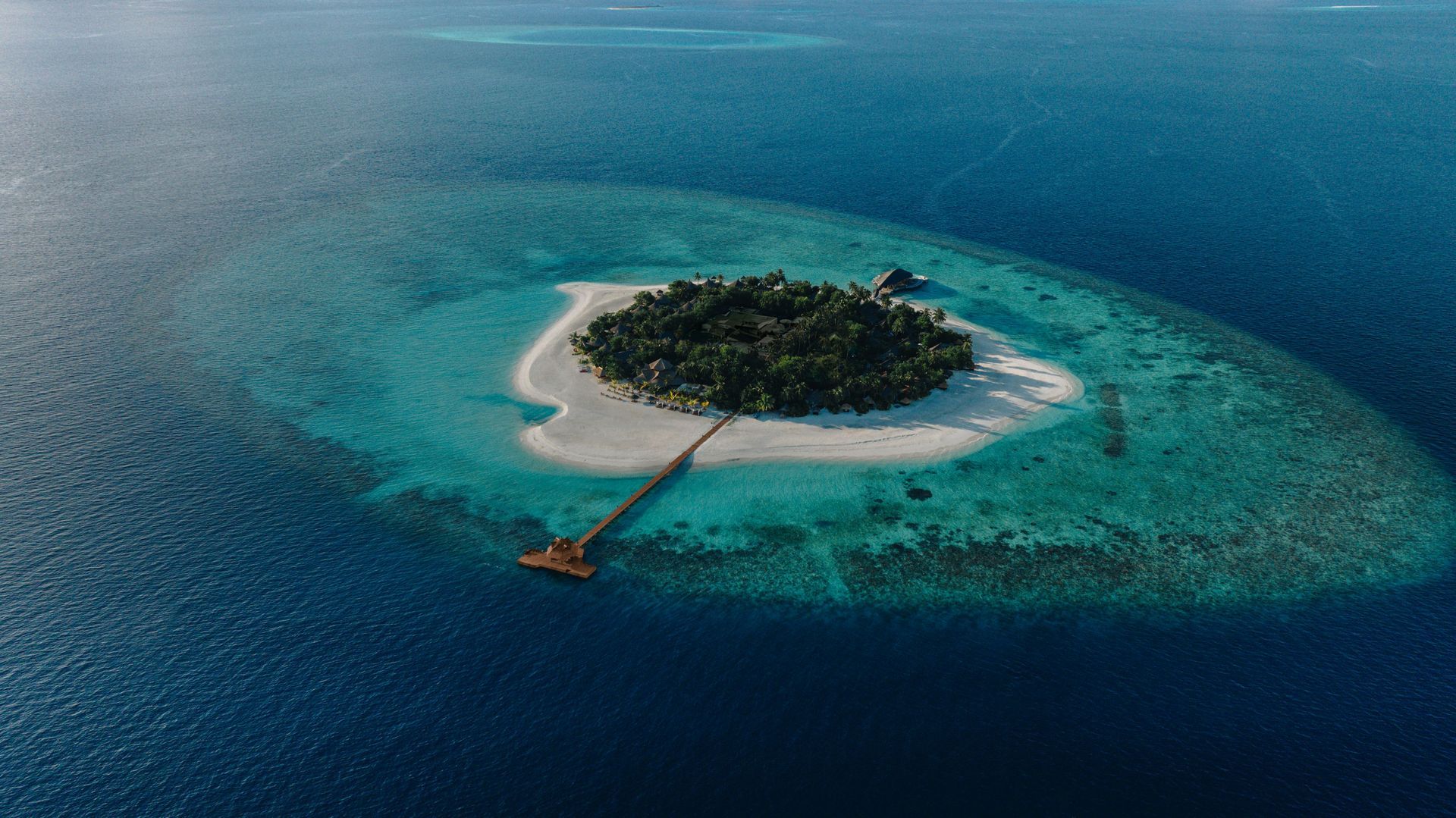 Aerial view of a small island with white sand, surrounded by turquoise and deep blue ocean. A pier extends from the island.