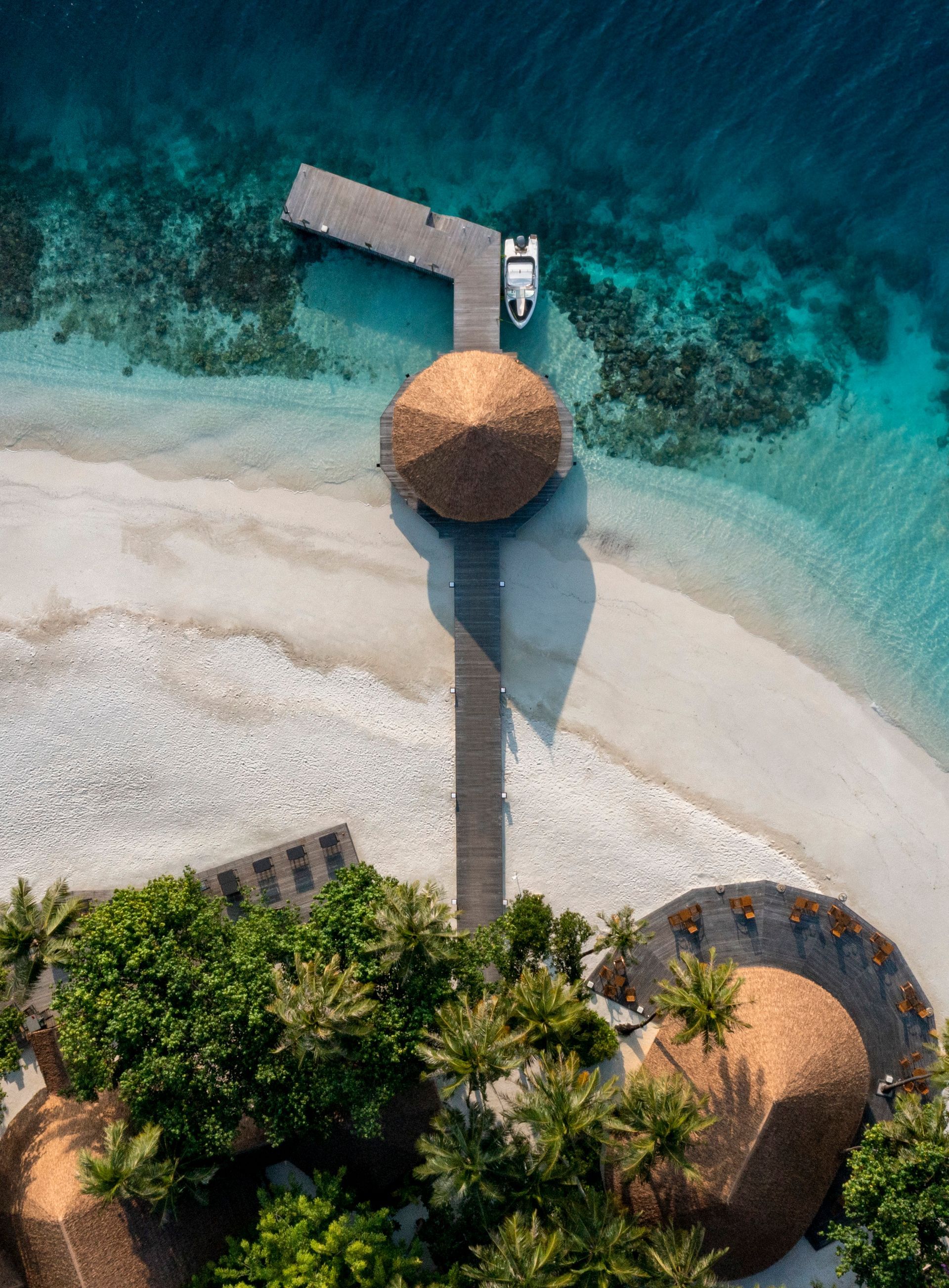 Aerial view: Beach resort with thatched-roof huts, pier, white sand, turquoise water, and greenery.
