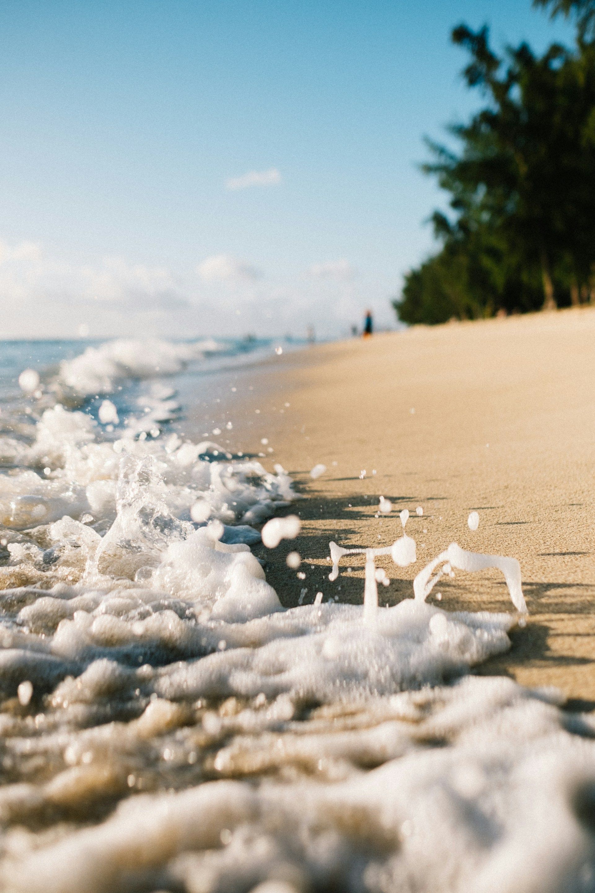 Waves crash on a sandy beach, with trees in the background under a bright blue sky.