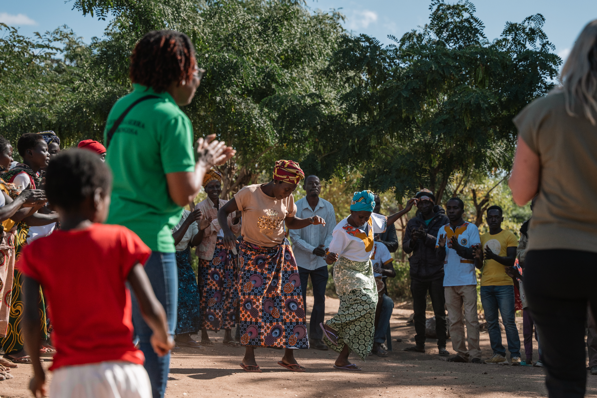 People dancing outdoors, surrounded by onlookers. A woman in green watches. Bright, sunny day.