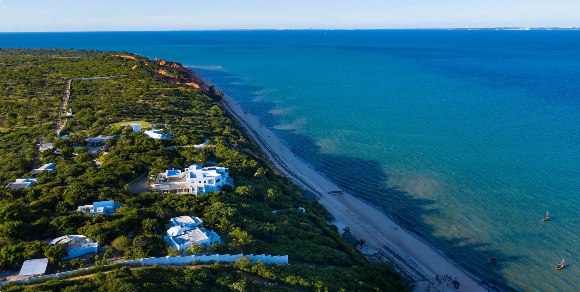 Aerial view of a coastline with white buildings, dense green trees, and a blue ocean under a bright sky.