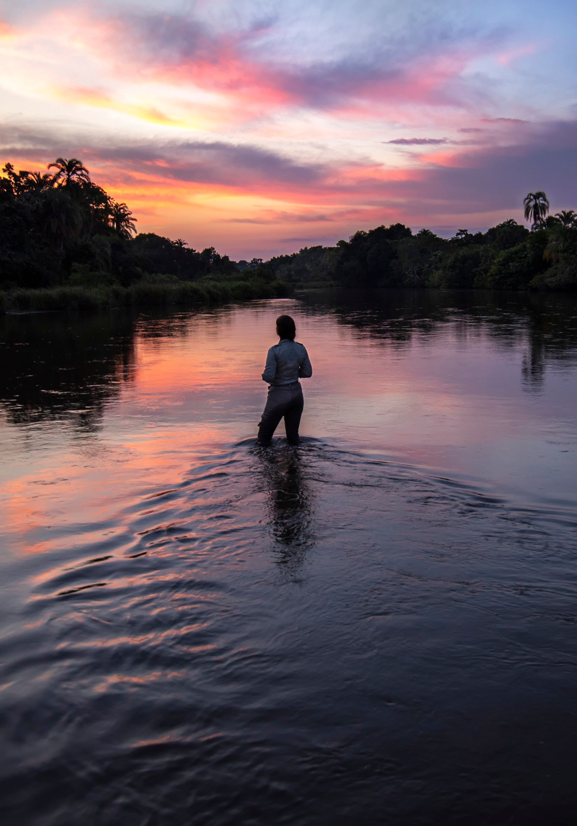 Person wading in a river at sunset; pink and orange sky reflects on the water.
