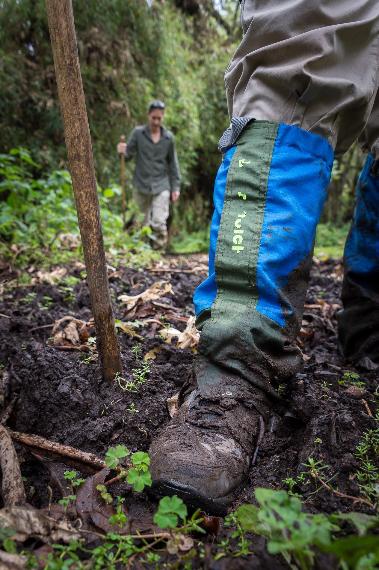 A hiker in mud, wearing blue and green leg gaiters, walks in a forest path, another person in the background.