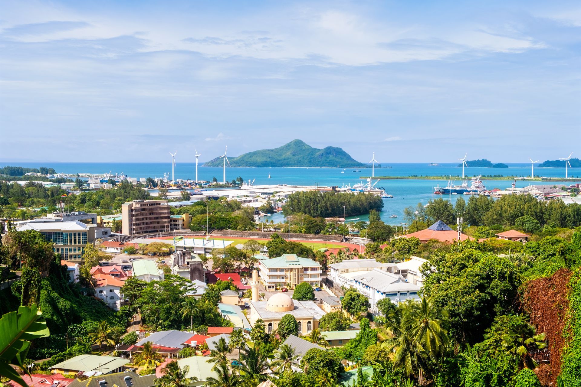Coastal city of Victoria, Seychelles, with buildings, harbor, and distant islands under a blue sky.