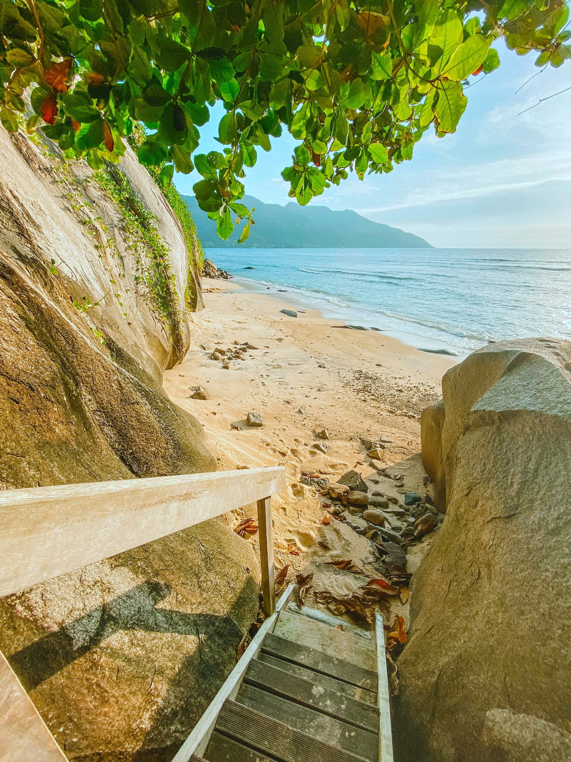 Wooden stairs lead to a sandy beach with blue water and a mountain in the distance; shaded by green foliage.