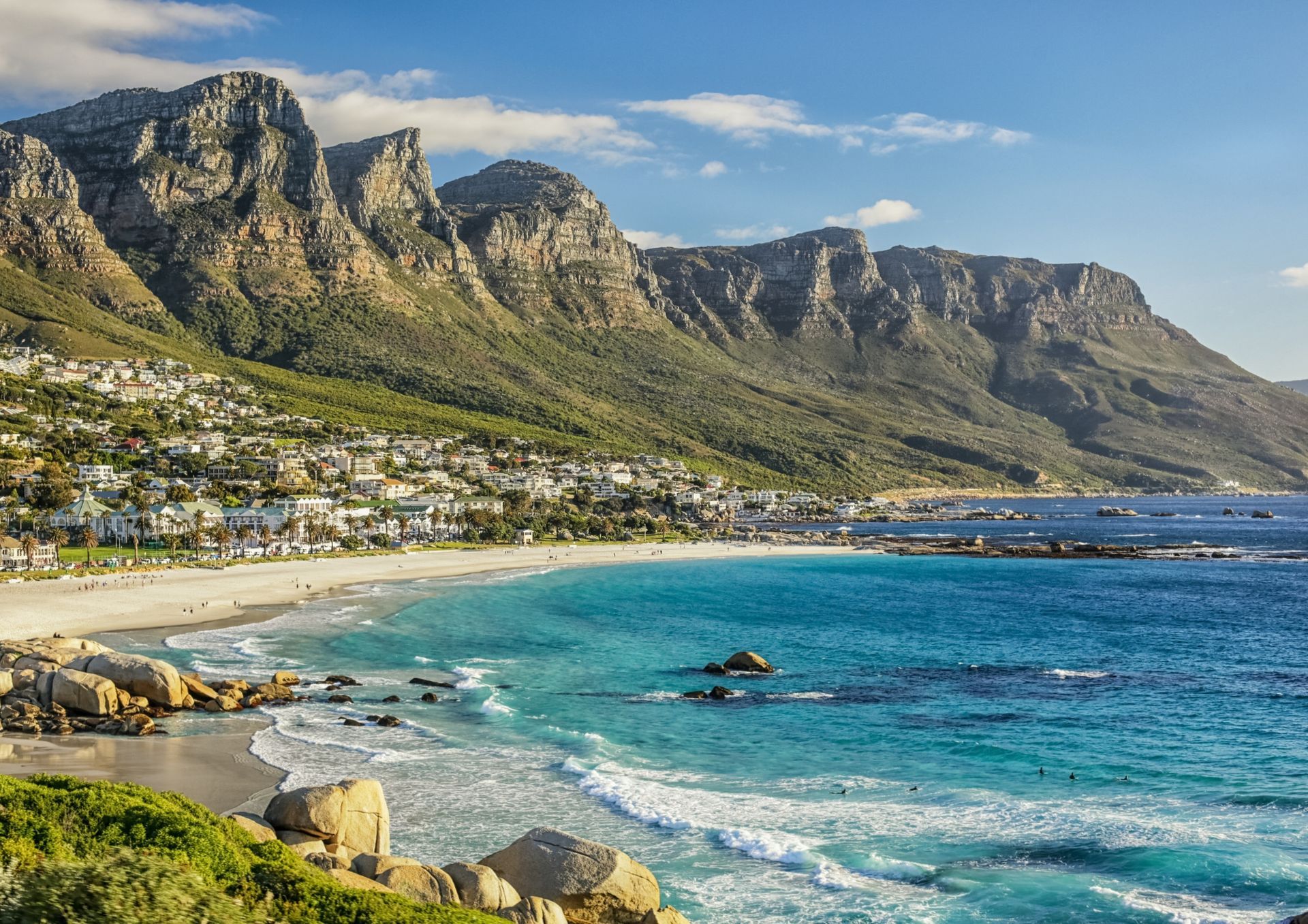 Beach with blue water, white sand, and a town backed by mountains under a blue sky.