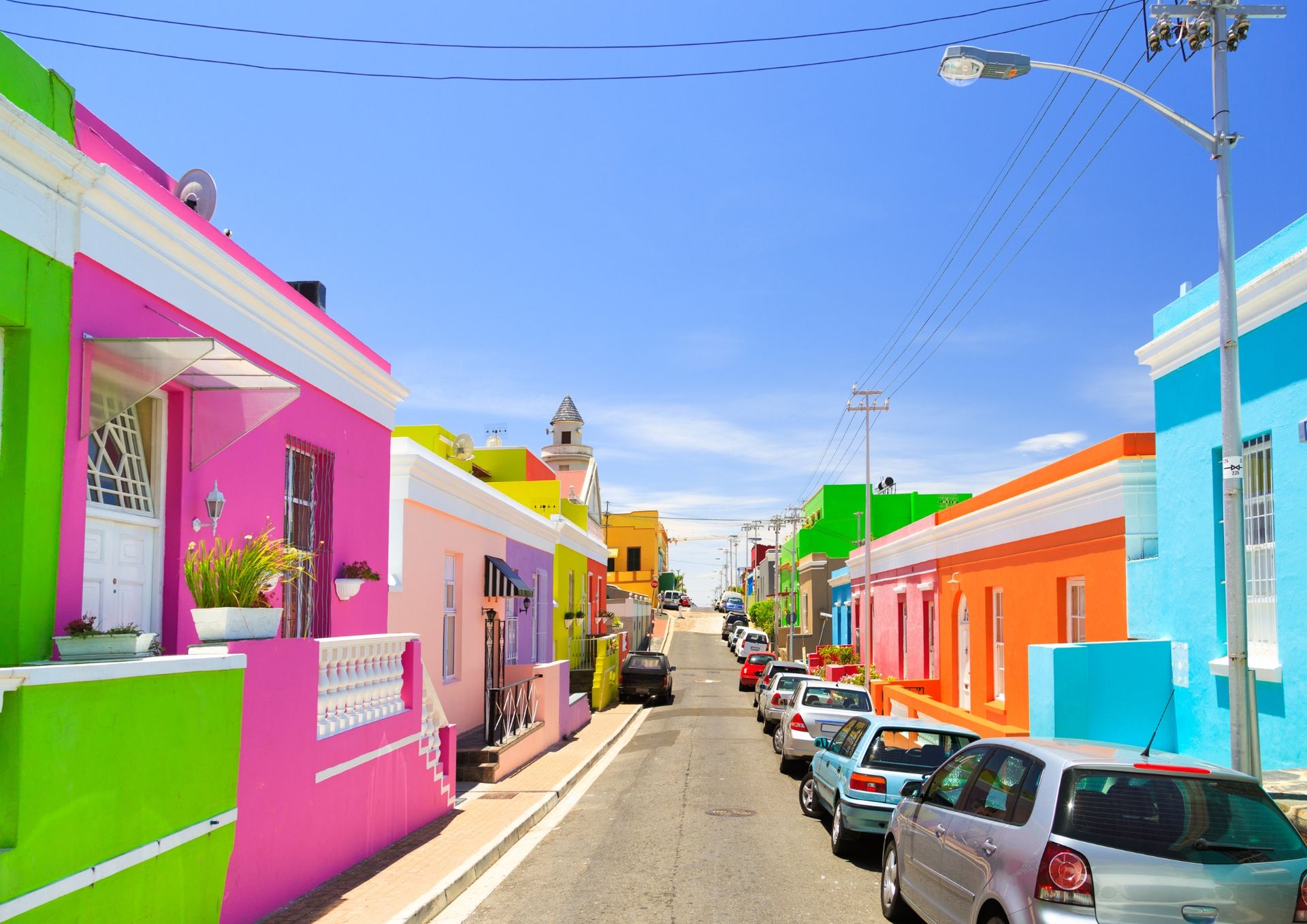 Colorful houses line a street in Bo-Kaap, Cape Town, South Africa, under a bright blue sky. Cars are parked along the road.