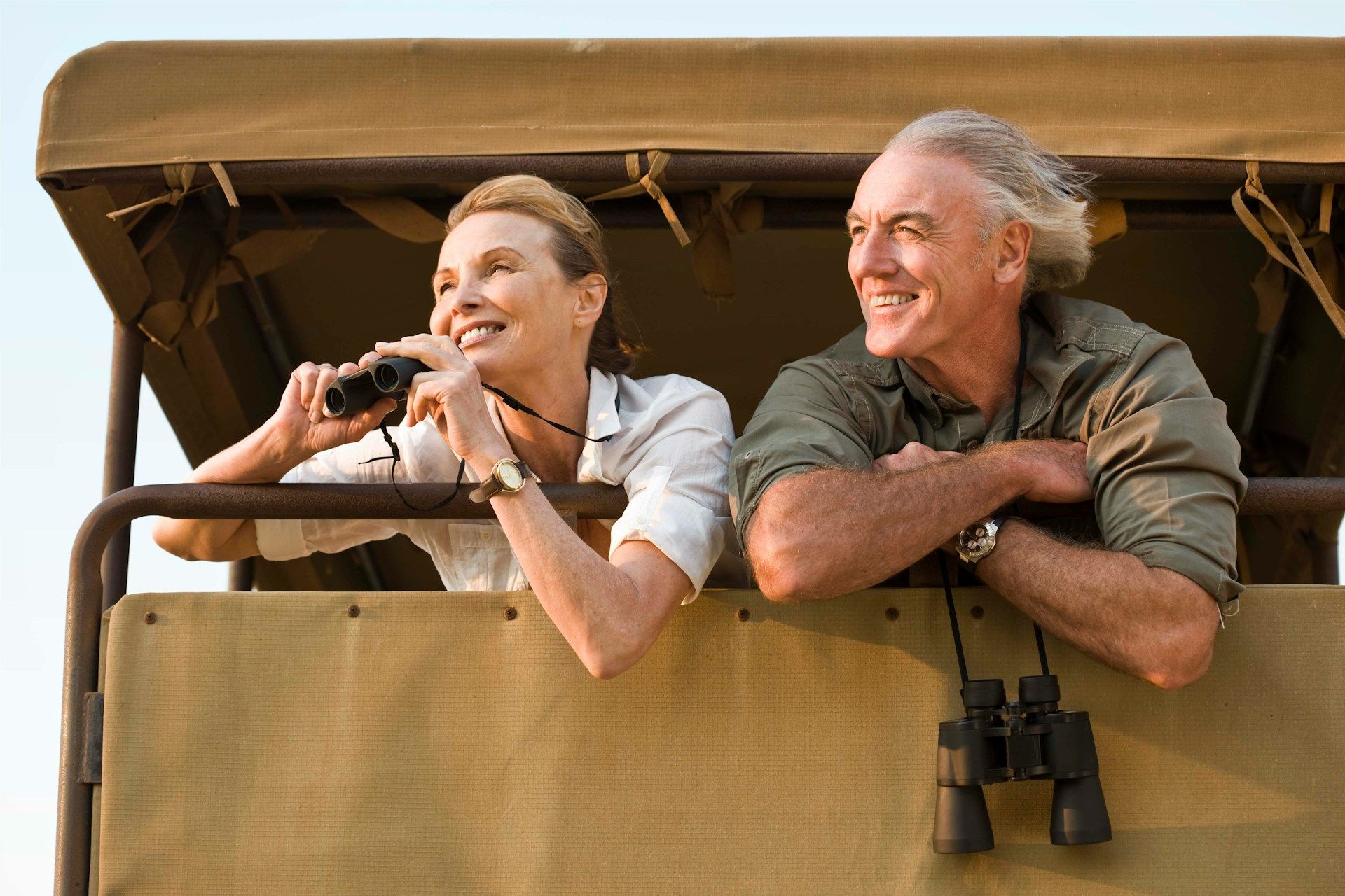 Older couple on safari, woman using binoculars, both smiling, in a vehicle.