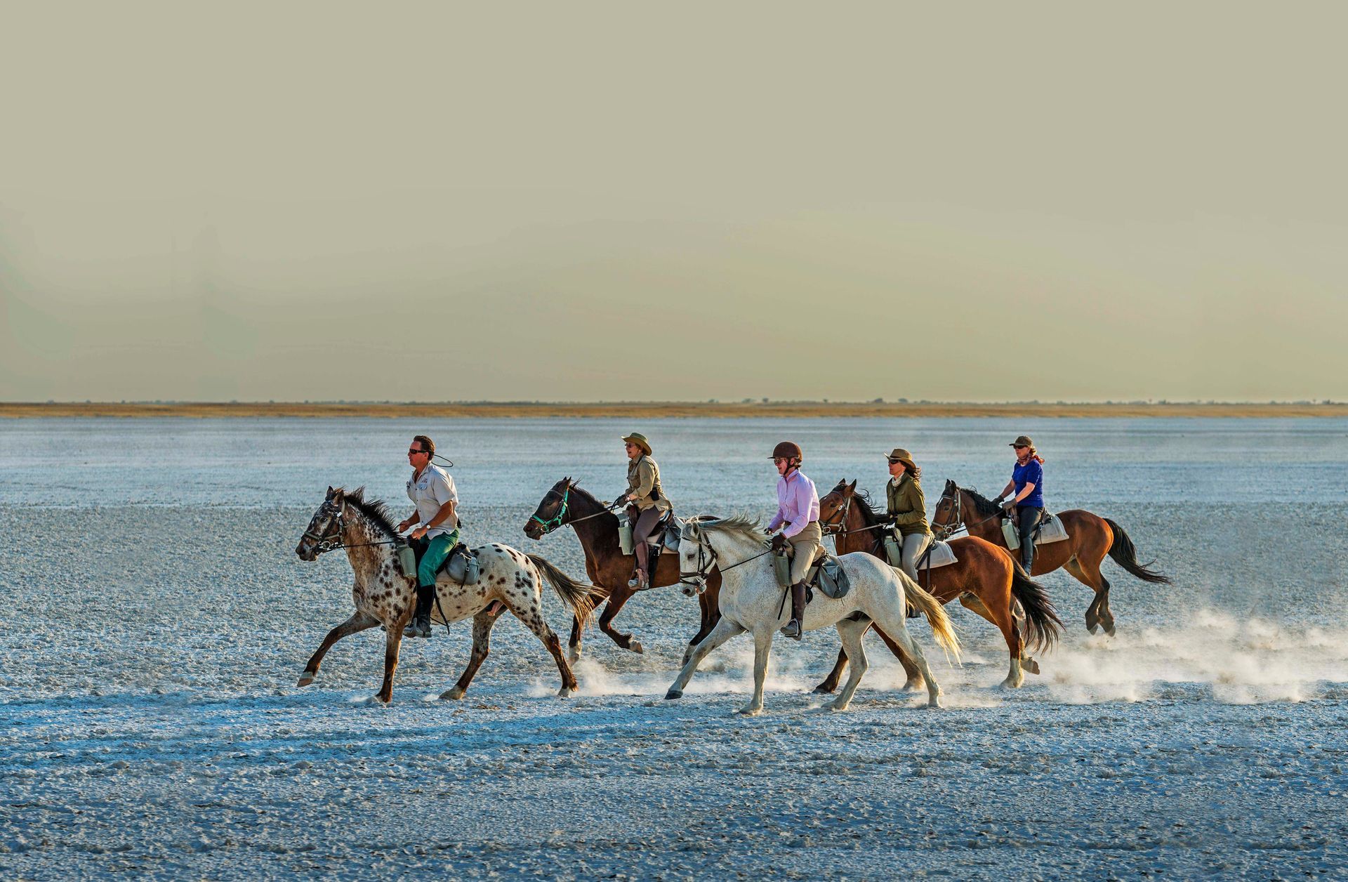 People riding horses across a vast, flat, light-colored plain under a hazy sky.