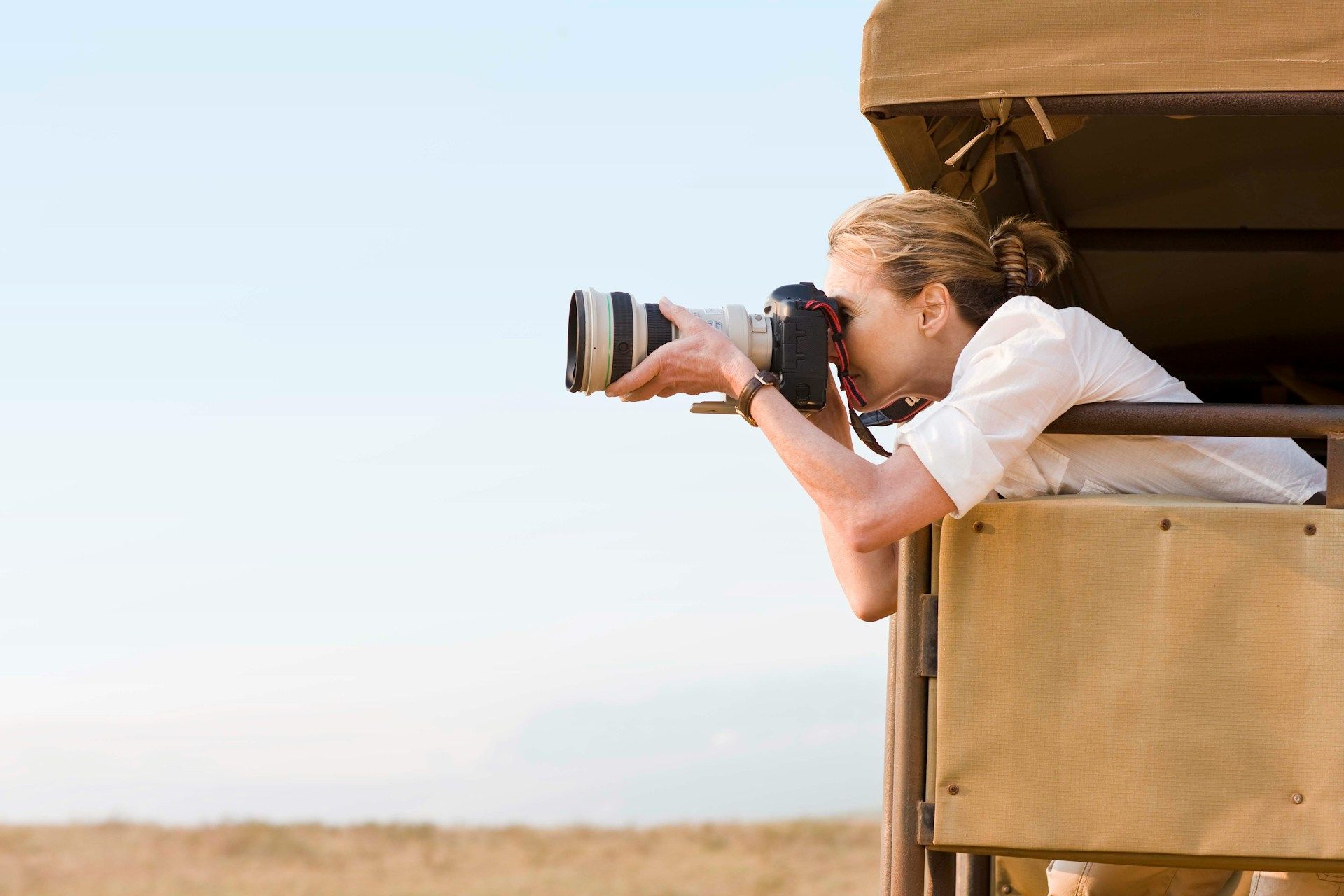 Woman taking photos from a safari vehicle, aiming camera at horizon. Blonde hair, pale shirt, sunny setting.