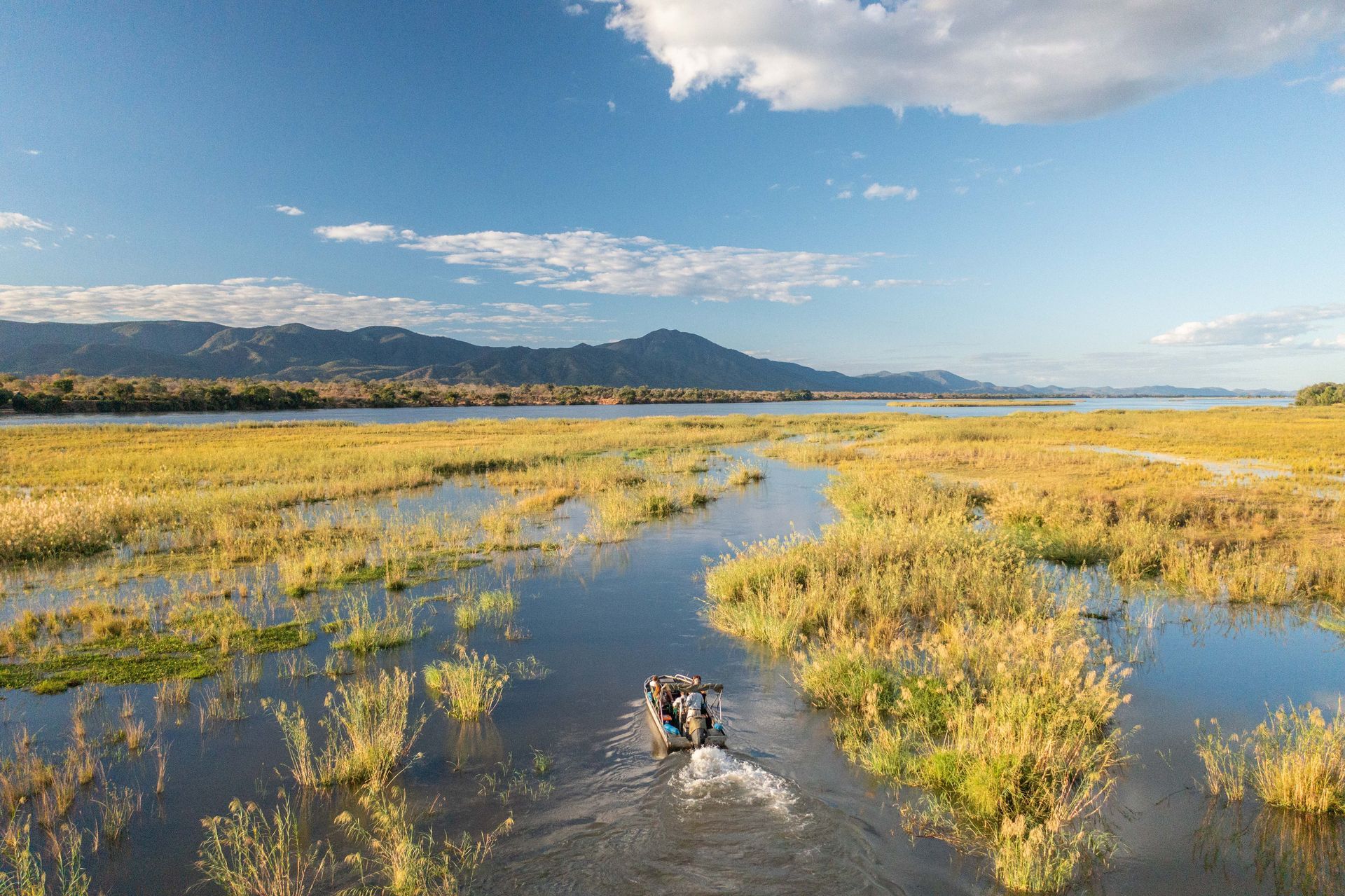 Two people in a boat navigate a flooded wetland, golden grasses and distant mountains under a blue sky.