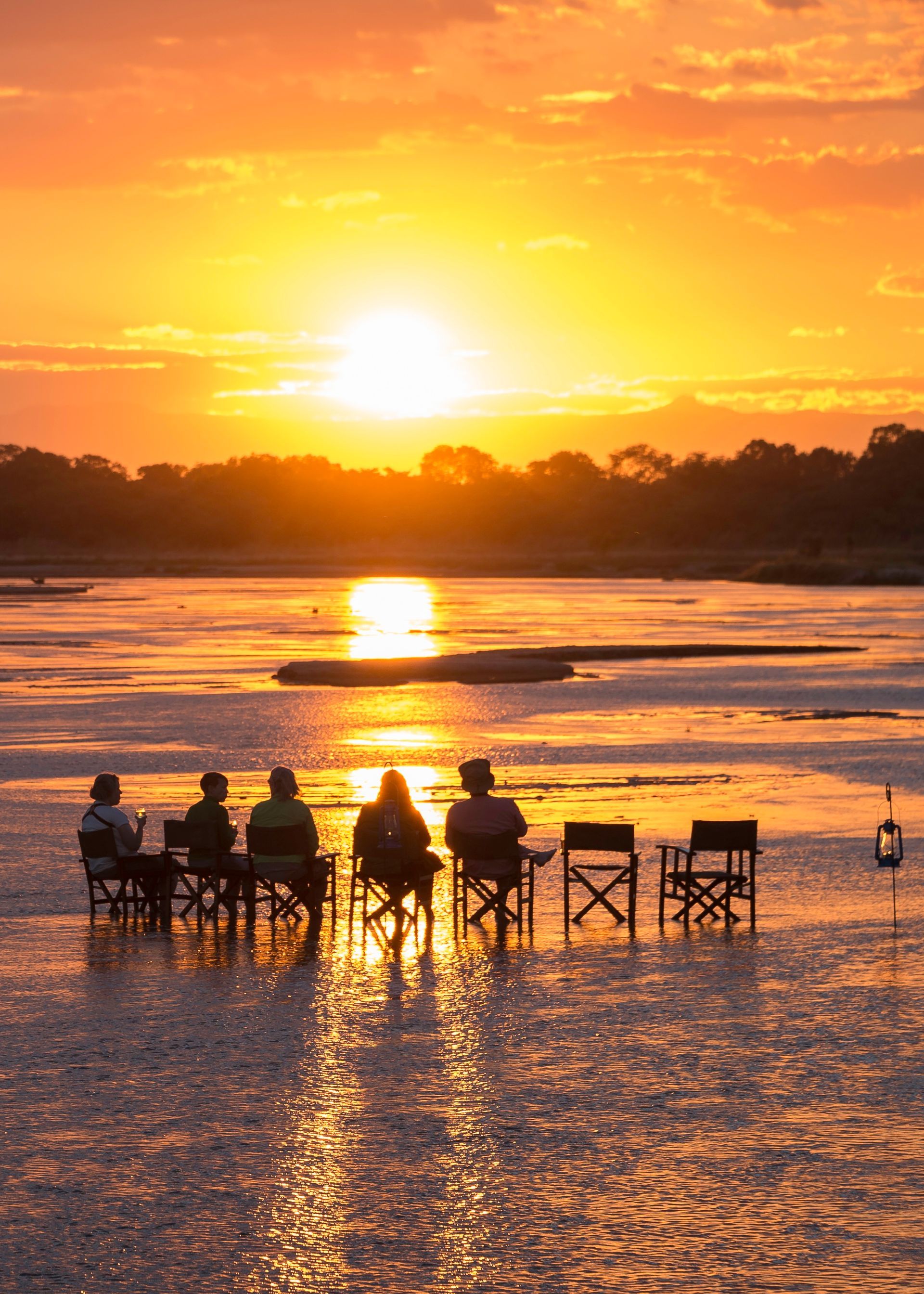 People seated in chairs watch a sunset over a river.