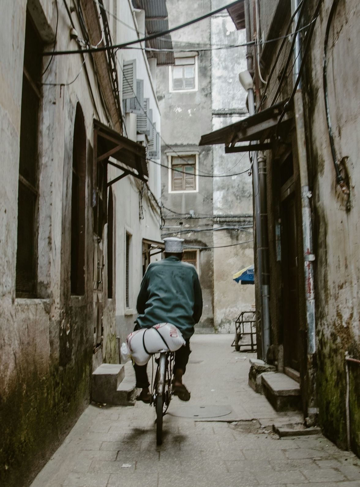 Man on bicycle in narrow alleyway, carrying white bundle, between aged buildings.