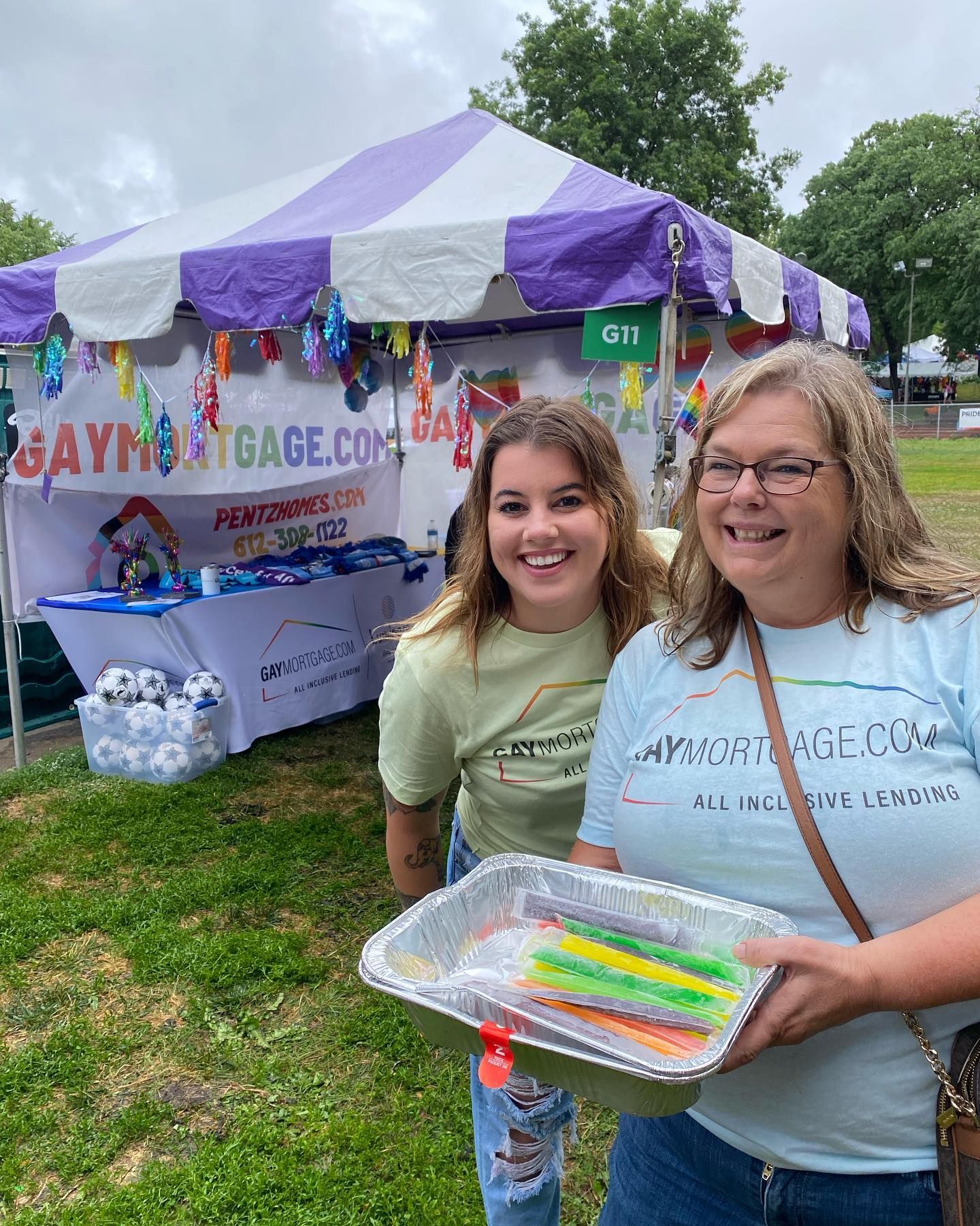Two women are standing in front of a purple and white tent holding a tray of candy.