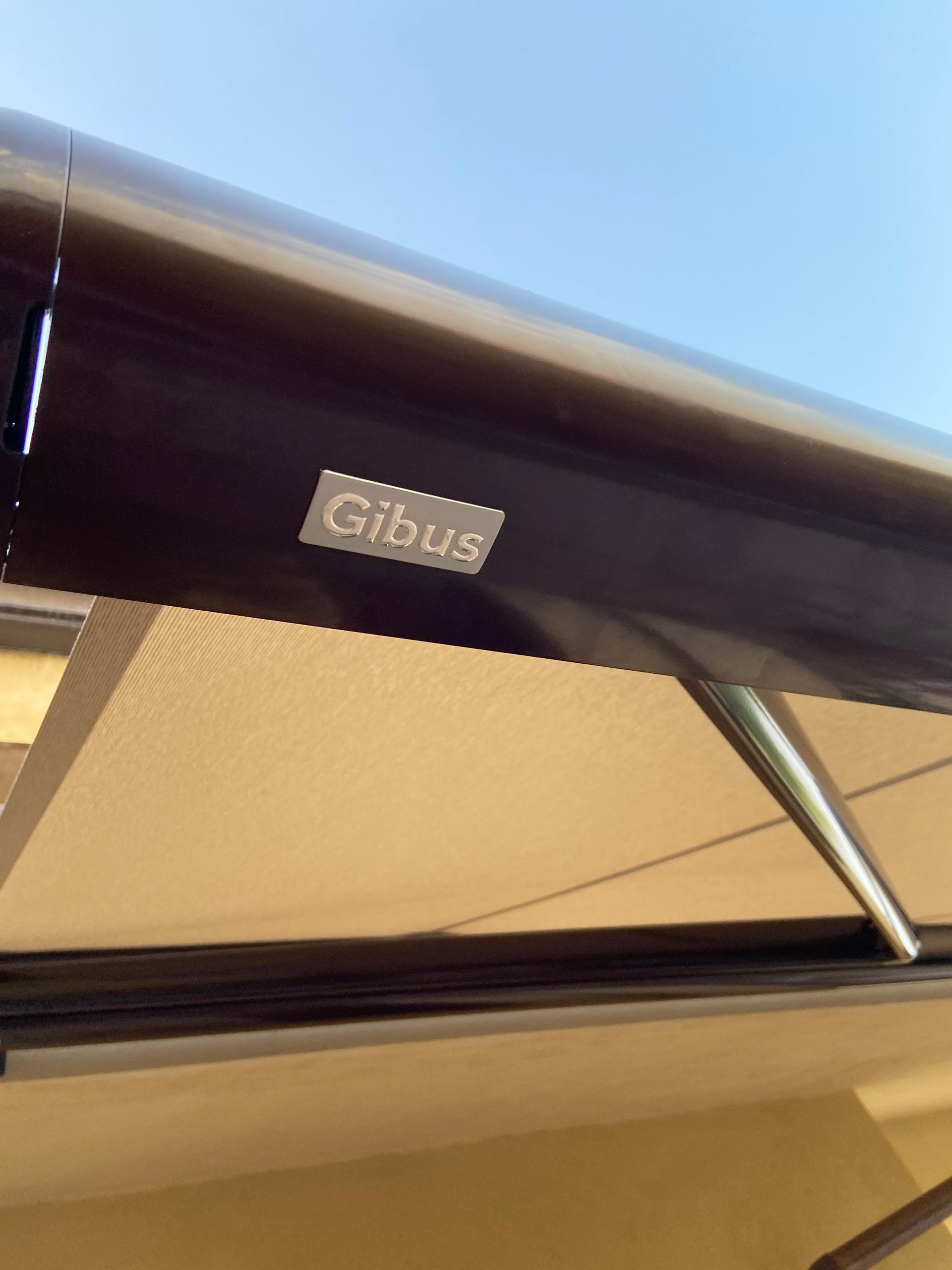 Primo piano di una tenda da sole Gibus, con telaio marrone scuro, tessuto beige e logo argentato sullo sfondo del cielo blu.