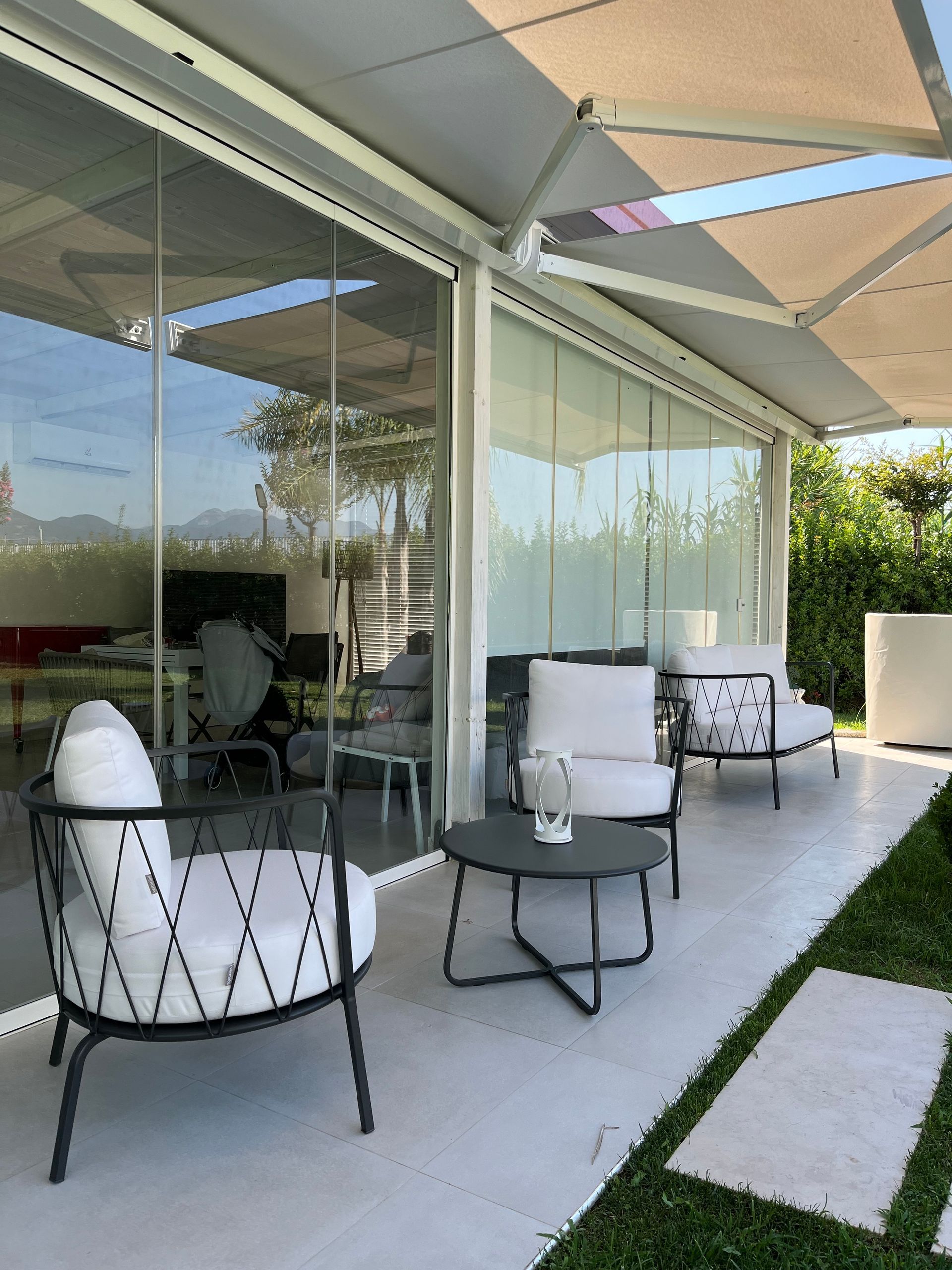 Area salotto all'aperto con pareti in vetro, tenda da sole e sedie con cuscini bianchi.