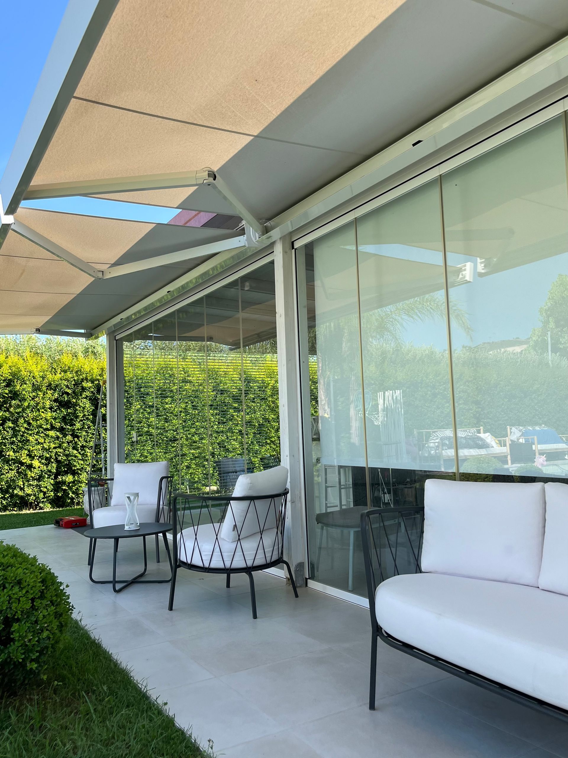 Patio con tenda da sole beige, pareti in vetro e mobili da esterno bianchi, vicino a una siepe.