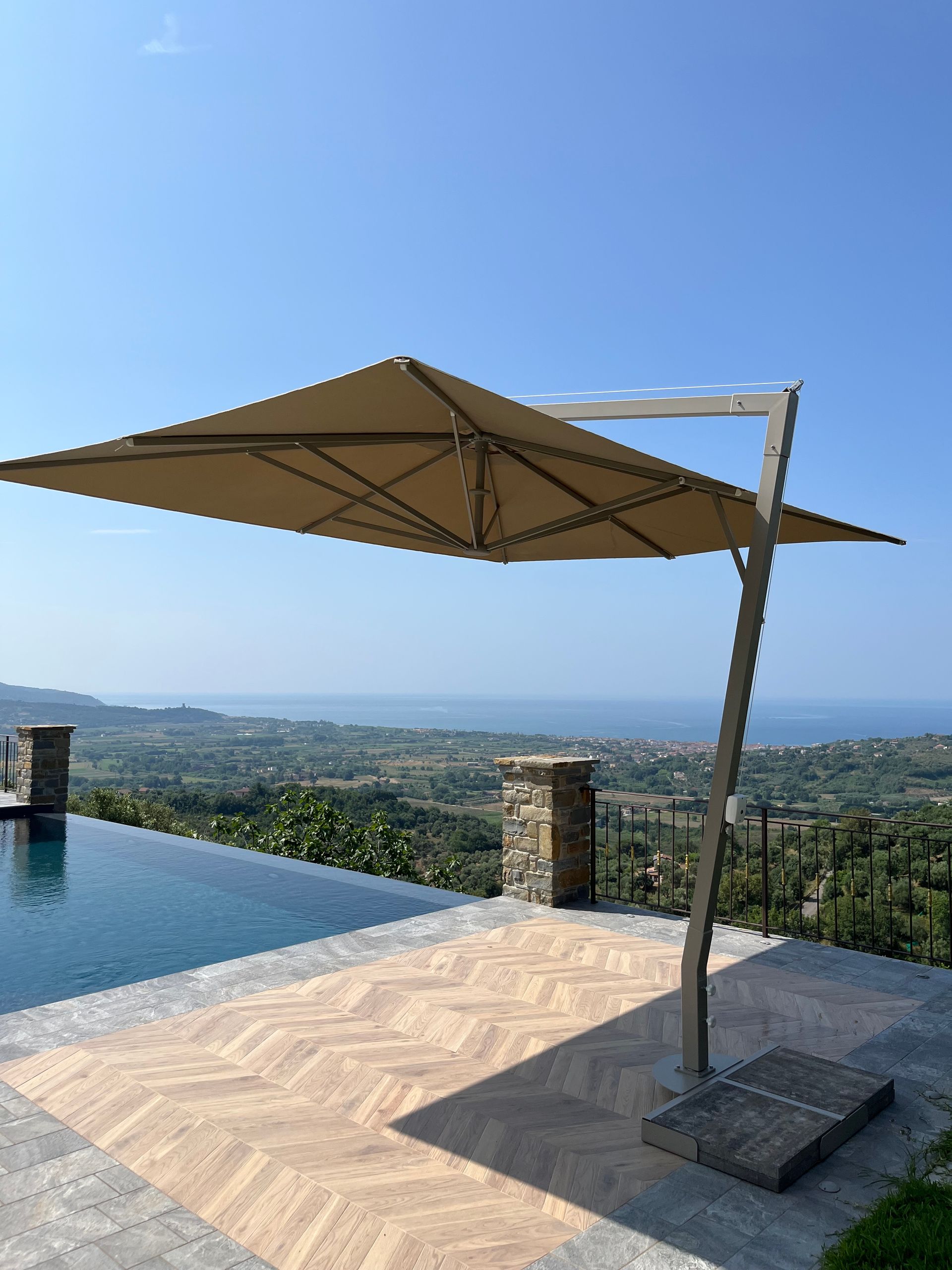 Ombrellone da patio beige a sbalzo sopra una piscina, con vista sul paesaggio e sul mare.