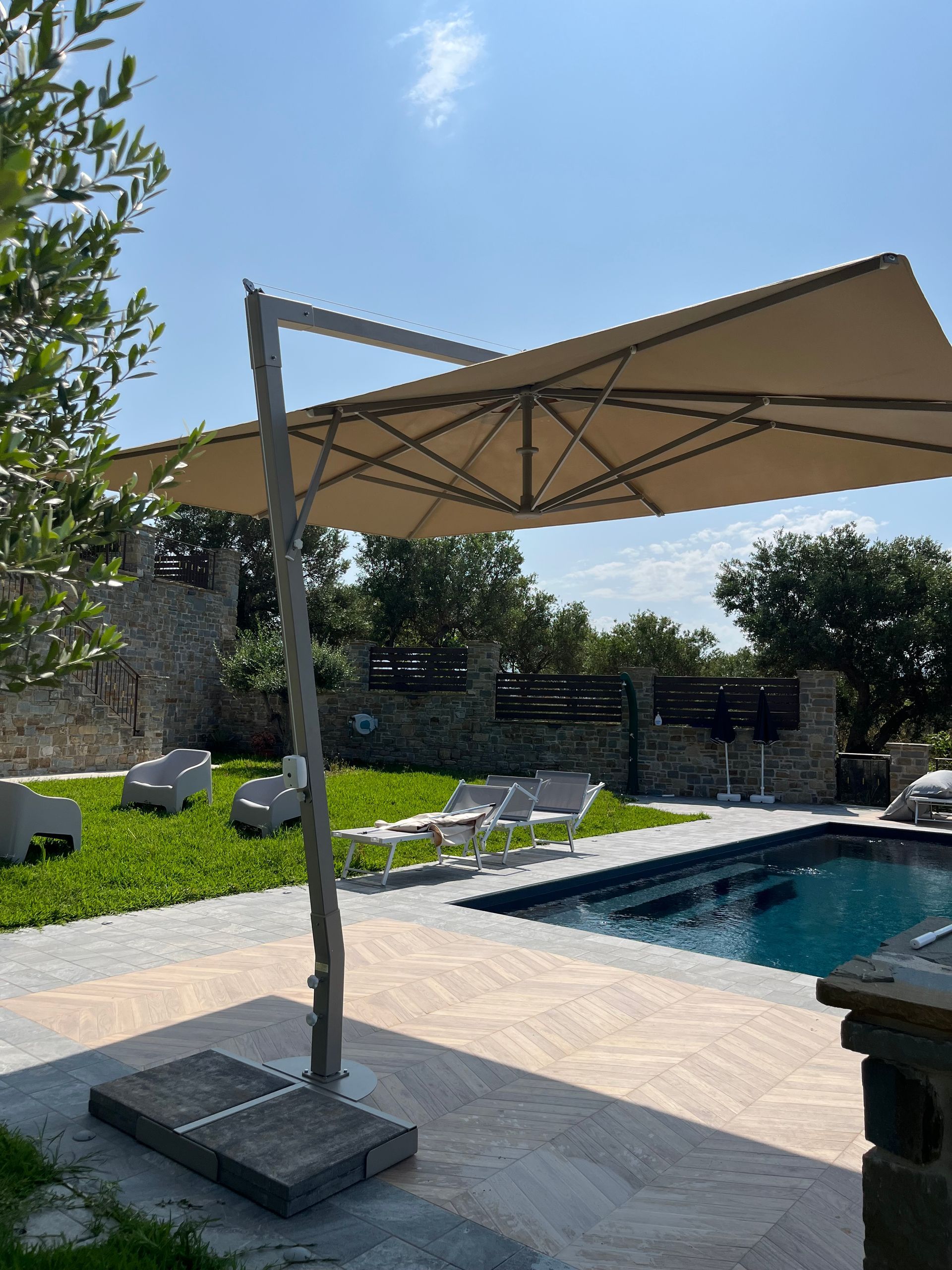 Grande ombrellone beige a sbalzo che ripara la piscina e le sedie a sdraio in una giornata di sole.