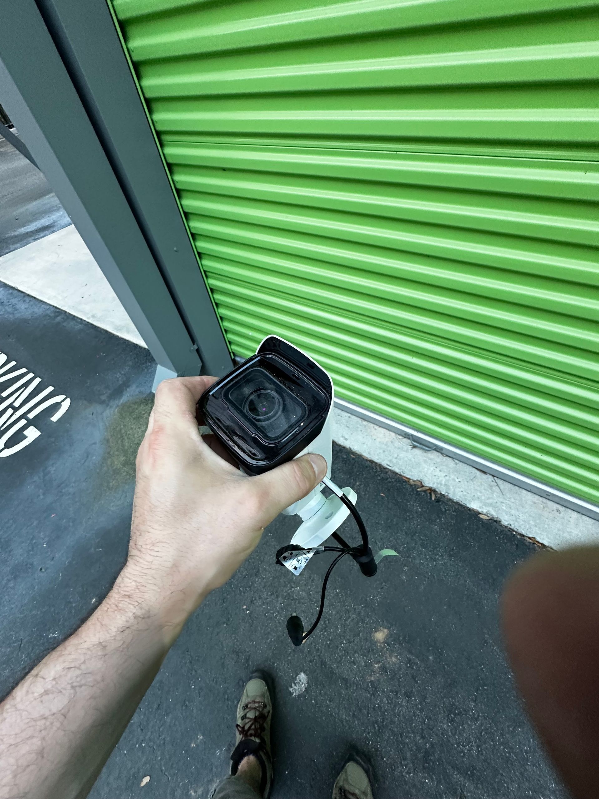 A person holds a black camera against a green storage unit door. The person's feet and arm are visible.