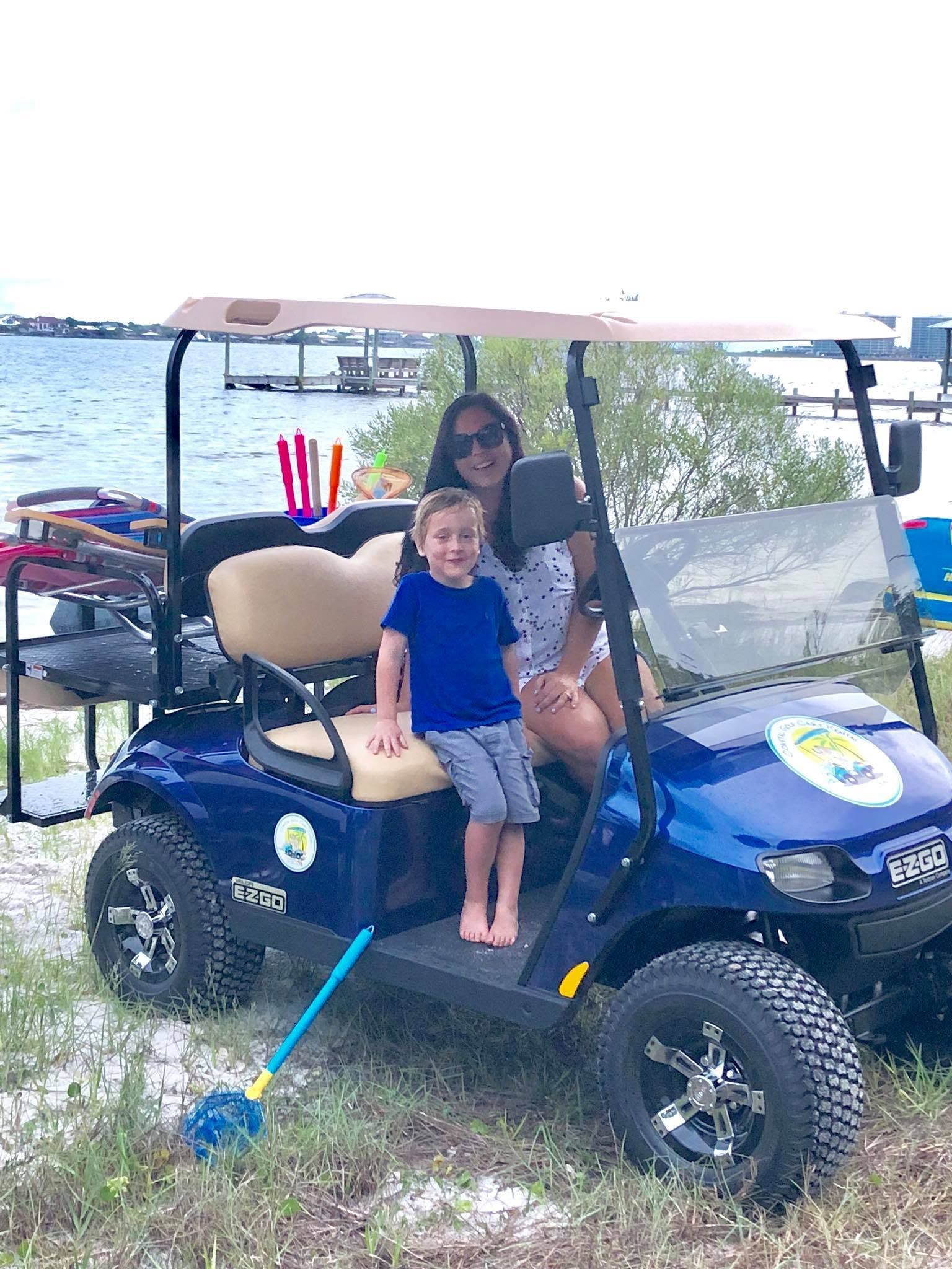 Two people seated in a blue golf cart by the water, with a fishing rod and tackle nearby.