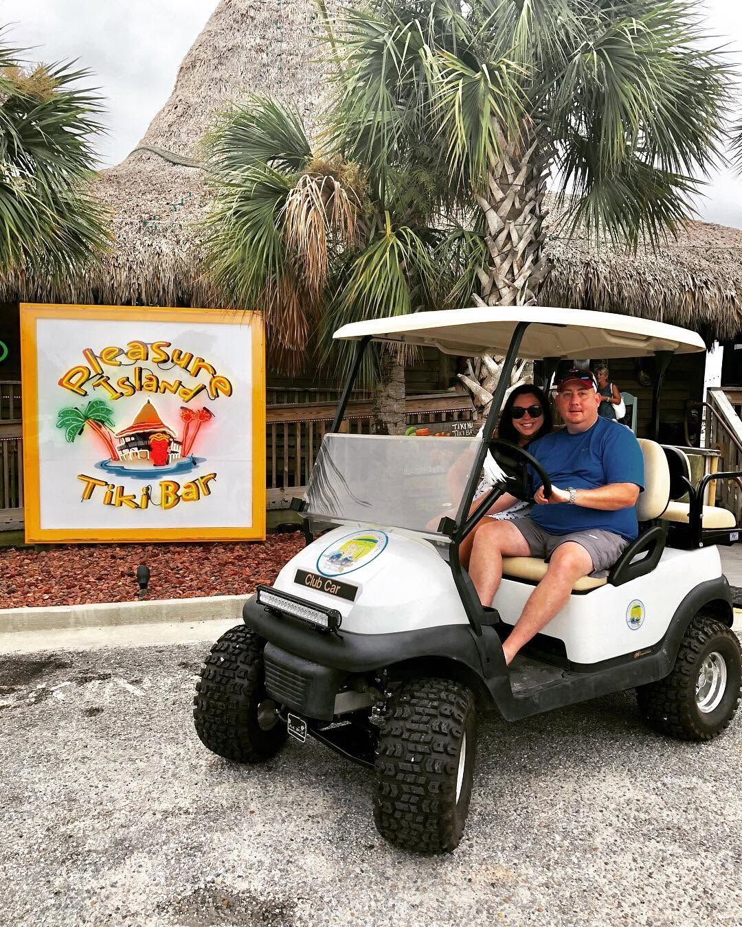 Two people in a golf cart beside a “Tiki Bar” sign in a tropical outdoor setting