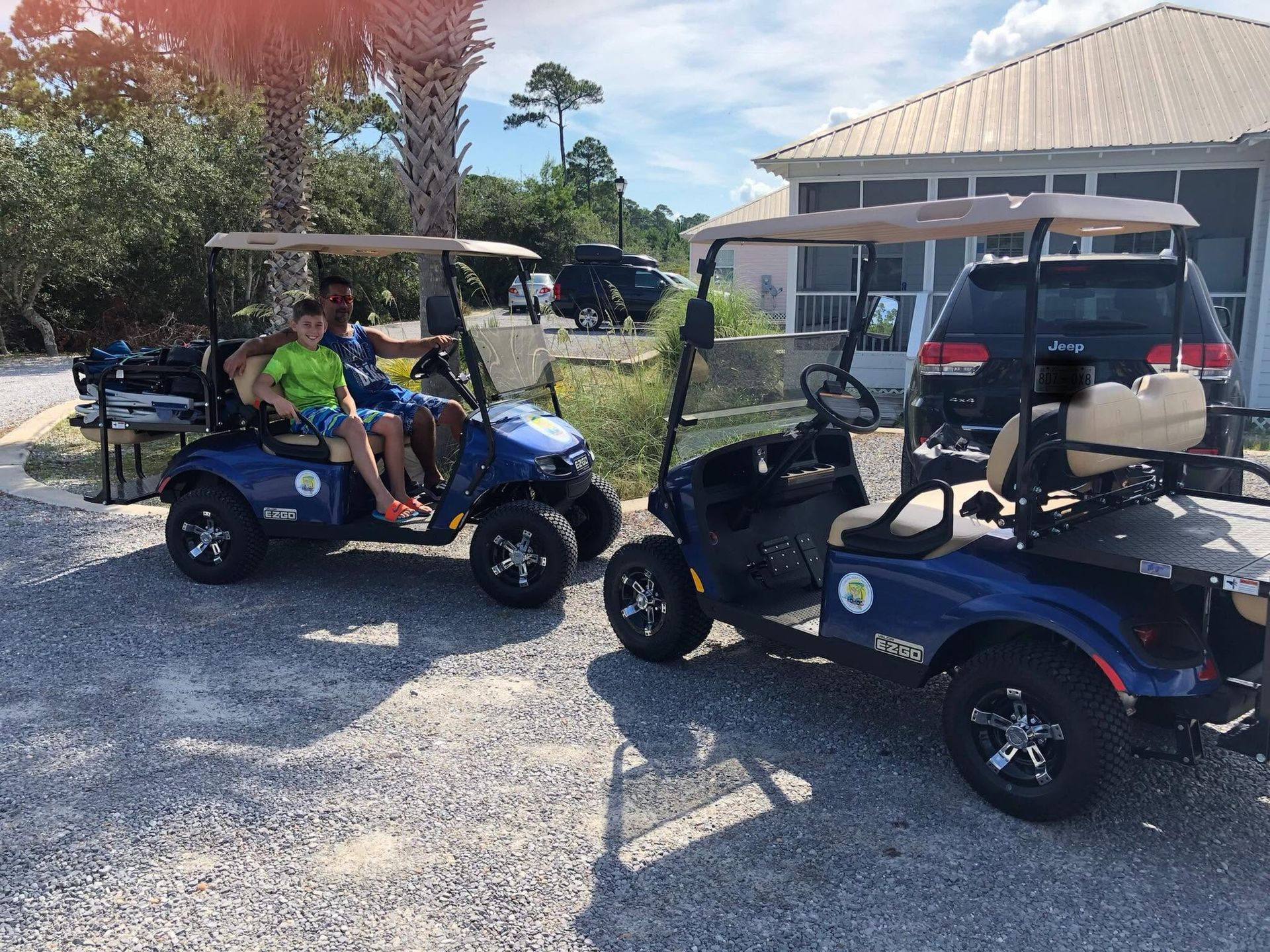 Two golf carts parked on a gravel lot, with people seated under canopies near a building.