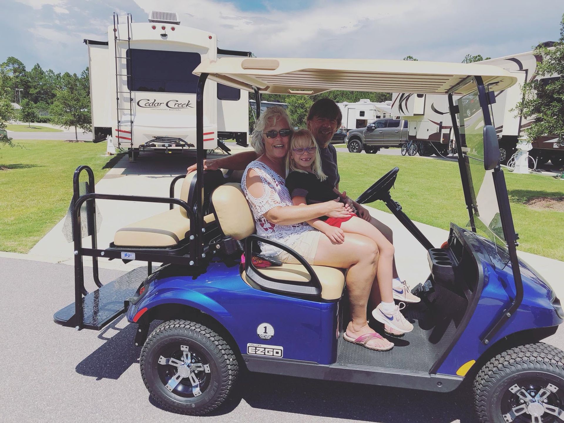 Two people sit in a blue golf cart on a grassy path under a white canopy.