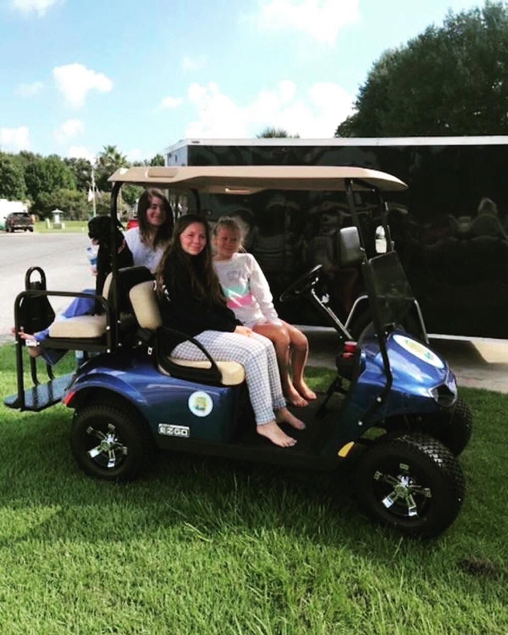 Two people sitting in a blue golf cart on a grassy field outdoors