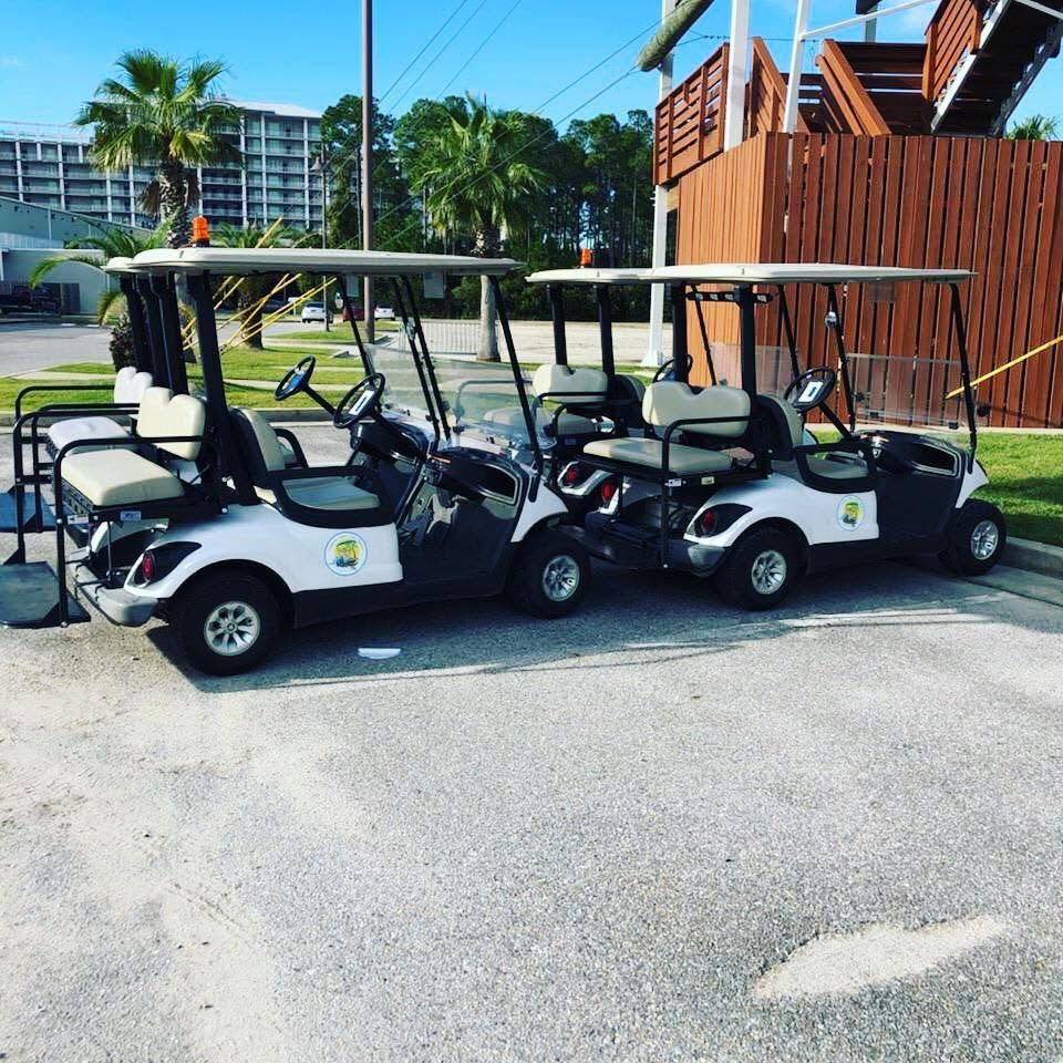 Three white golf carts parked side by side on a sunny street near a wooden fence.