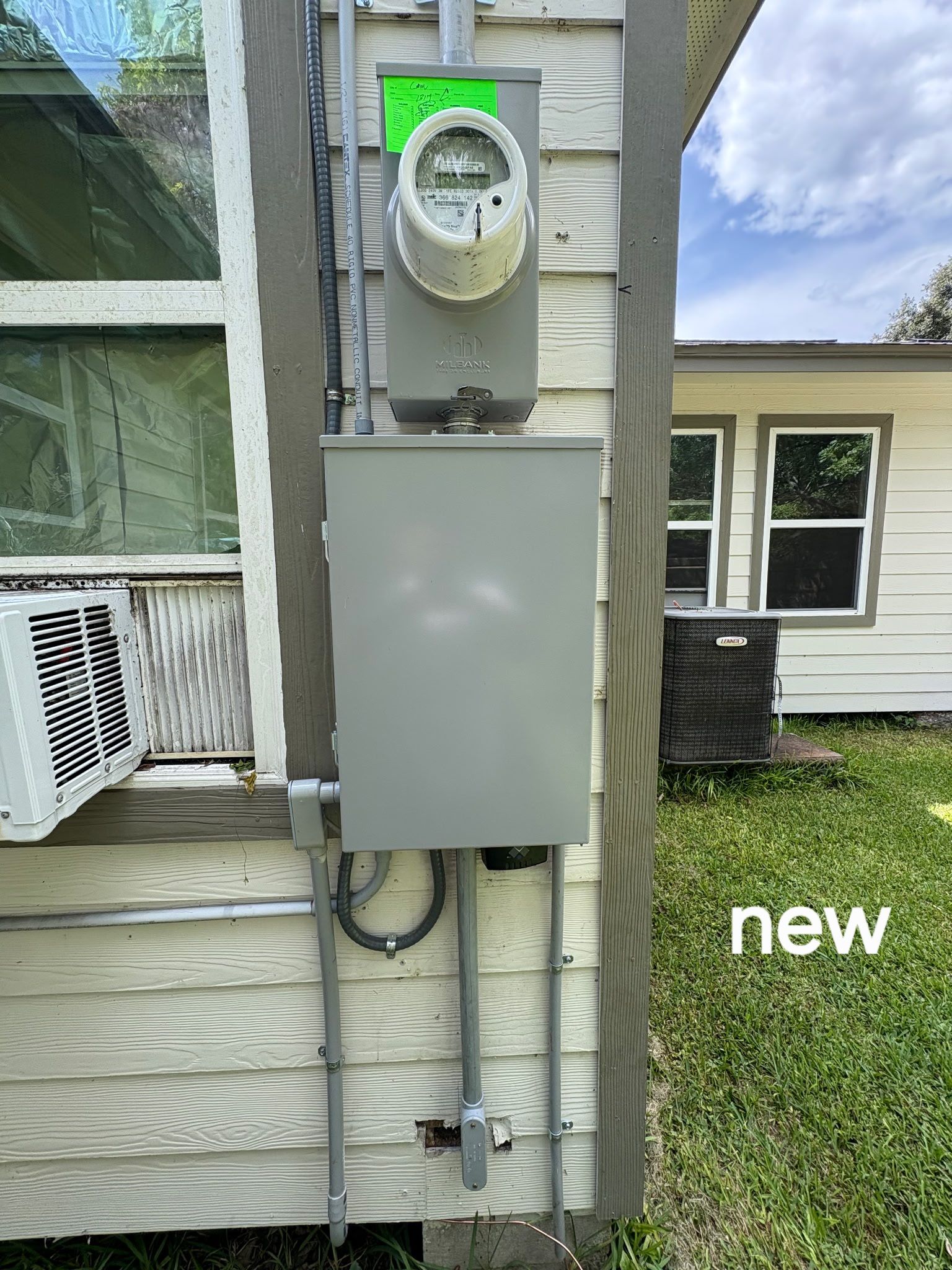 Outdoor electrical meter and breaker box mounted on house siding beside a window air conditioner.