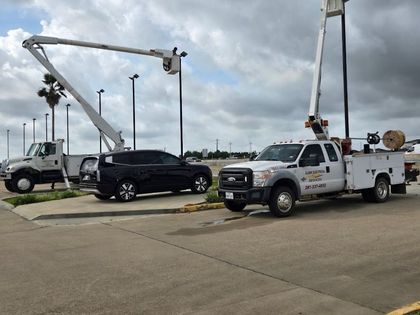 Two utility trucks with bucket lifts parked beside a black SUV on a cloudy roadside.