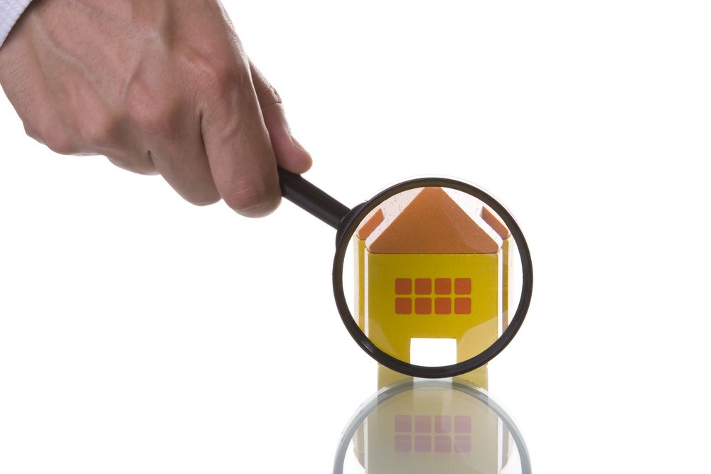 A hand holding a magnifying glass focuses on a small, yellow toy house, highlighting close scrutiny or inspection.